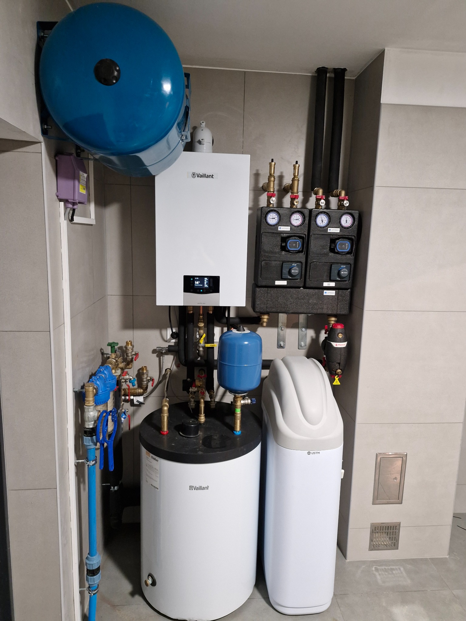 Kompletna instalacja grzewcza Vaillant z zasobnikiem, zmiękczaczem wody USTM i grupą pompową Afriso, zainstalowana w schludnym pomieszczeniu z szarymi płytkami.