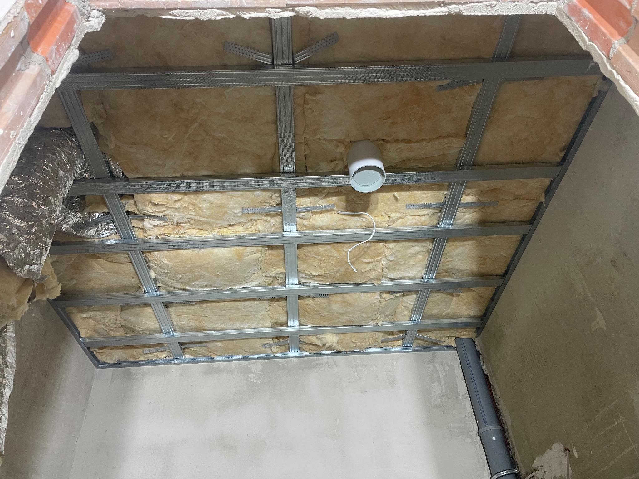 Metalowa konstrukcja sufitu podwieszanego z izolacją z wełny mineralnej, widoczny fragment instalacji wentylacyjnej i rura kanalizacyjna w rogu pomieszczenia.