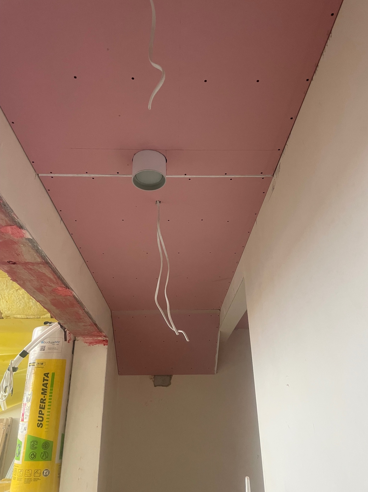 Różowa płyta karton gips na suficie z zamontowanym oświetleniem punktowym i wystającymi kablami. Widoczne łączenia płyt i otwory na wkręty.