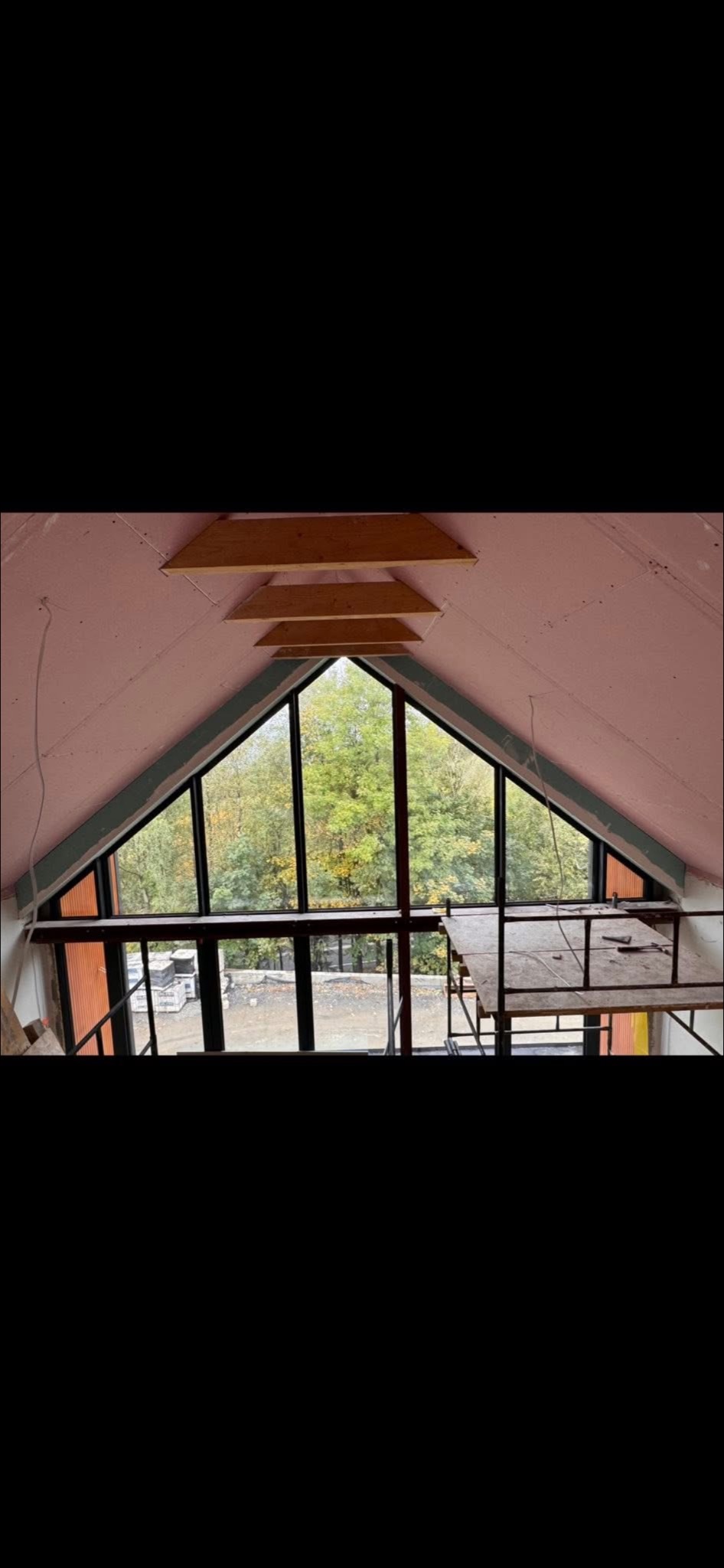 Poddasze z oknem w kształcie trójkąta, widok na drzewa. Ściany wykończone płytami gipsowymi, widoczne drewniane elementy konstrukcyjne i rusztowanie. Prace wykończeniowe.