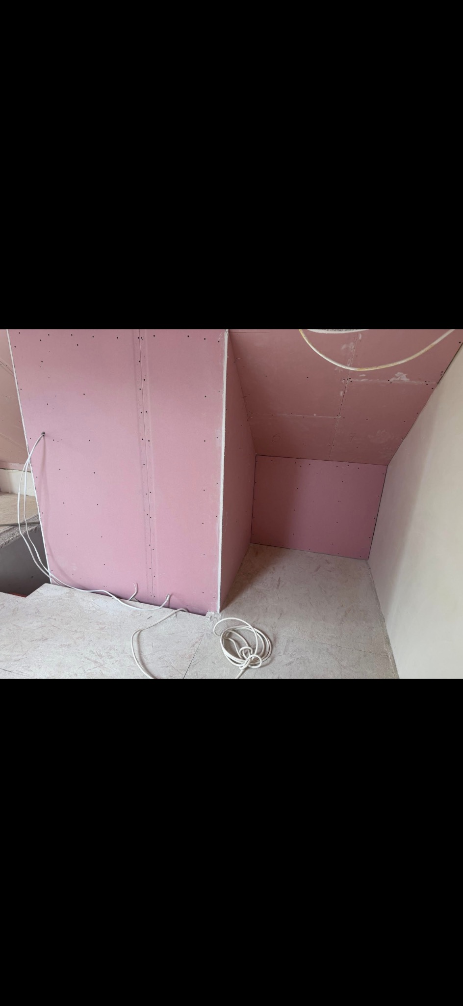 Różowe płyty kartonowo-gipsowe zamontowane na ścianach i suficie w rogu pomieszczenia. Widoczne kable elektryczne i surowa podłoga. Prace wykończeniowe w trakcie.