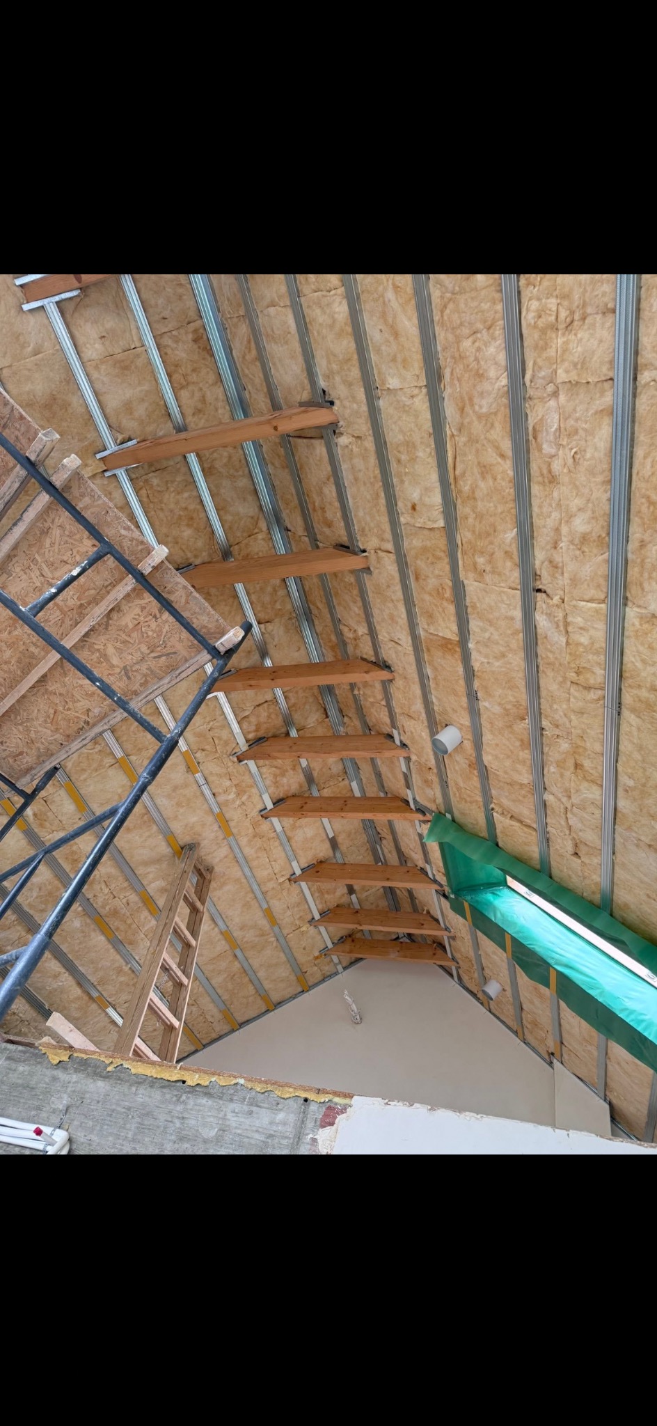 Skośny strych z izolacją z wełny mineralnej, metalowymi profilami i drewnianymi elementami konstrukcyjnymi. Widoczne rusztowanie i drabina.