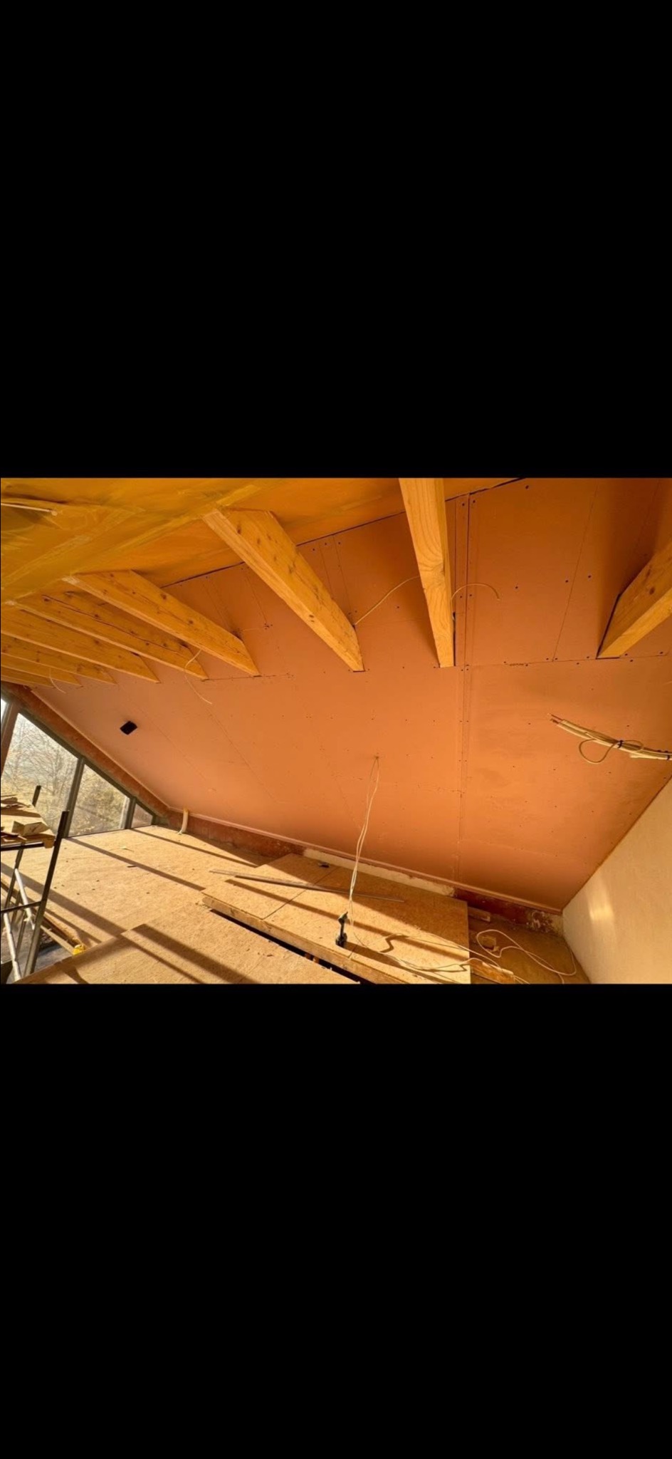 Skośny sufit z drewnianą konstrukcją i zamontowanymi płytami. Widoczne kable i fragment rusztowania. Ujęcie z dołu, w trakcie prac wykończeniowych.