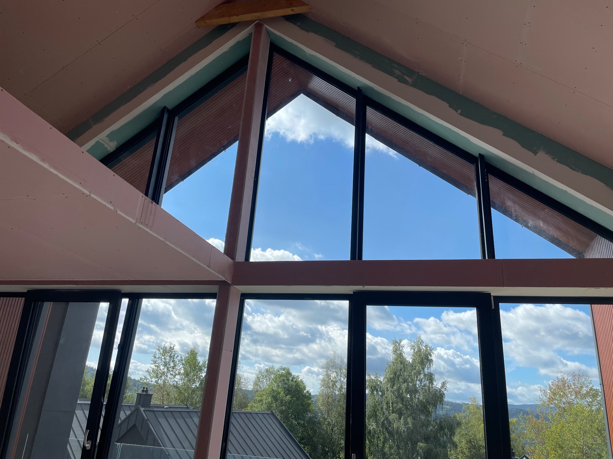 Montaż płyt gipsowo-kartonowych na poddaszu z oknami o nietypowym kształcie. Widoczne łączenia płyt i fragmenty szpachlowania. W tle błękitne niebo i zieleń drzew.