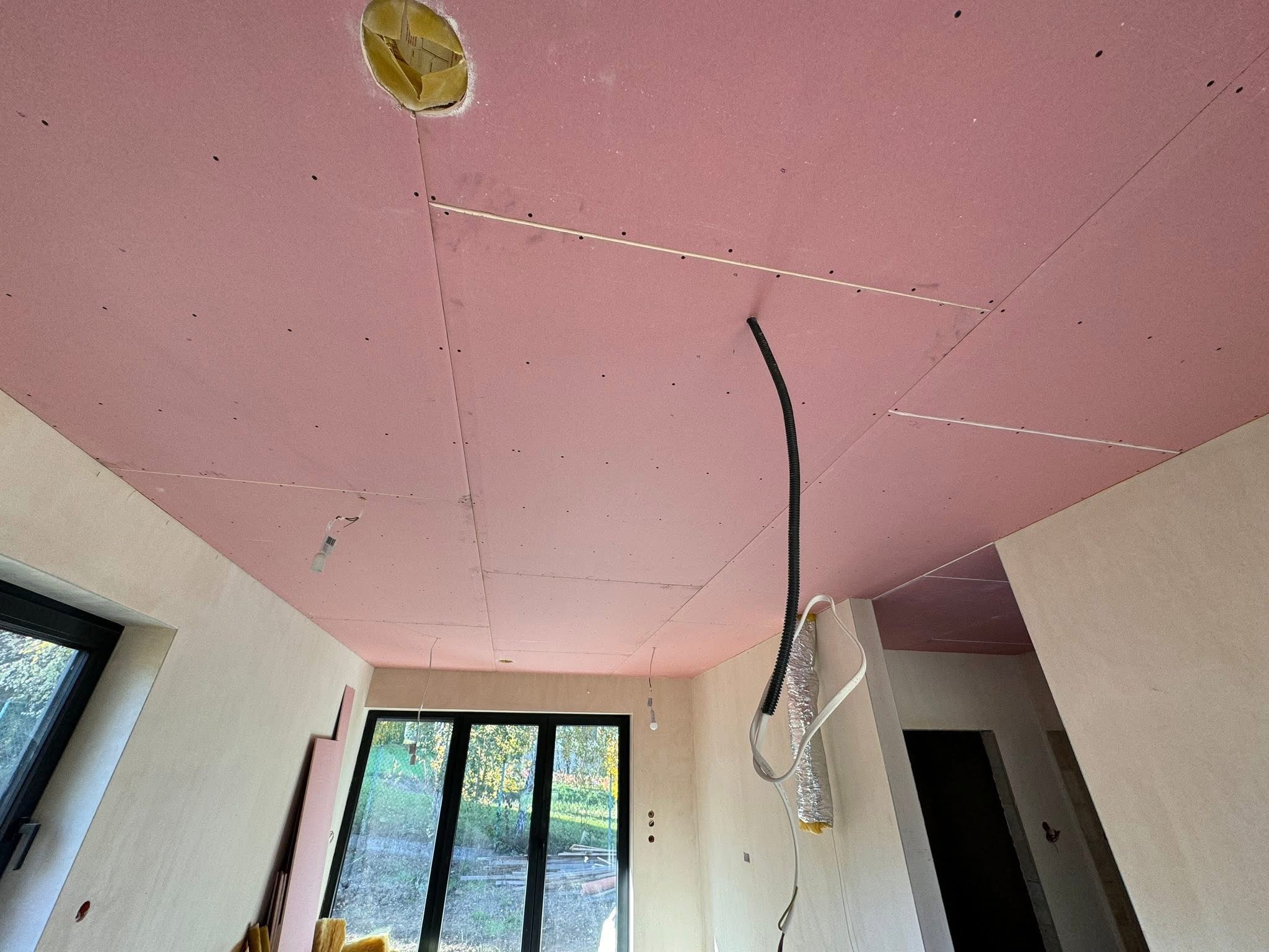 Różowa płyta karton-gips na suficie z widocznymi przewodami i otworem na oświetlenie, w trakcie montażu w jasnym wnętrzu z oknami.