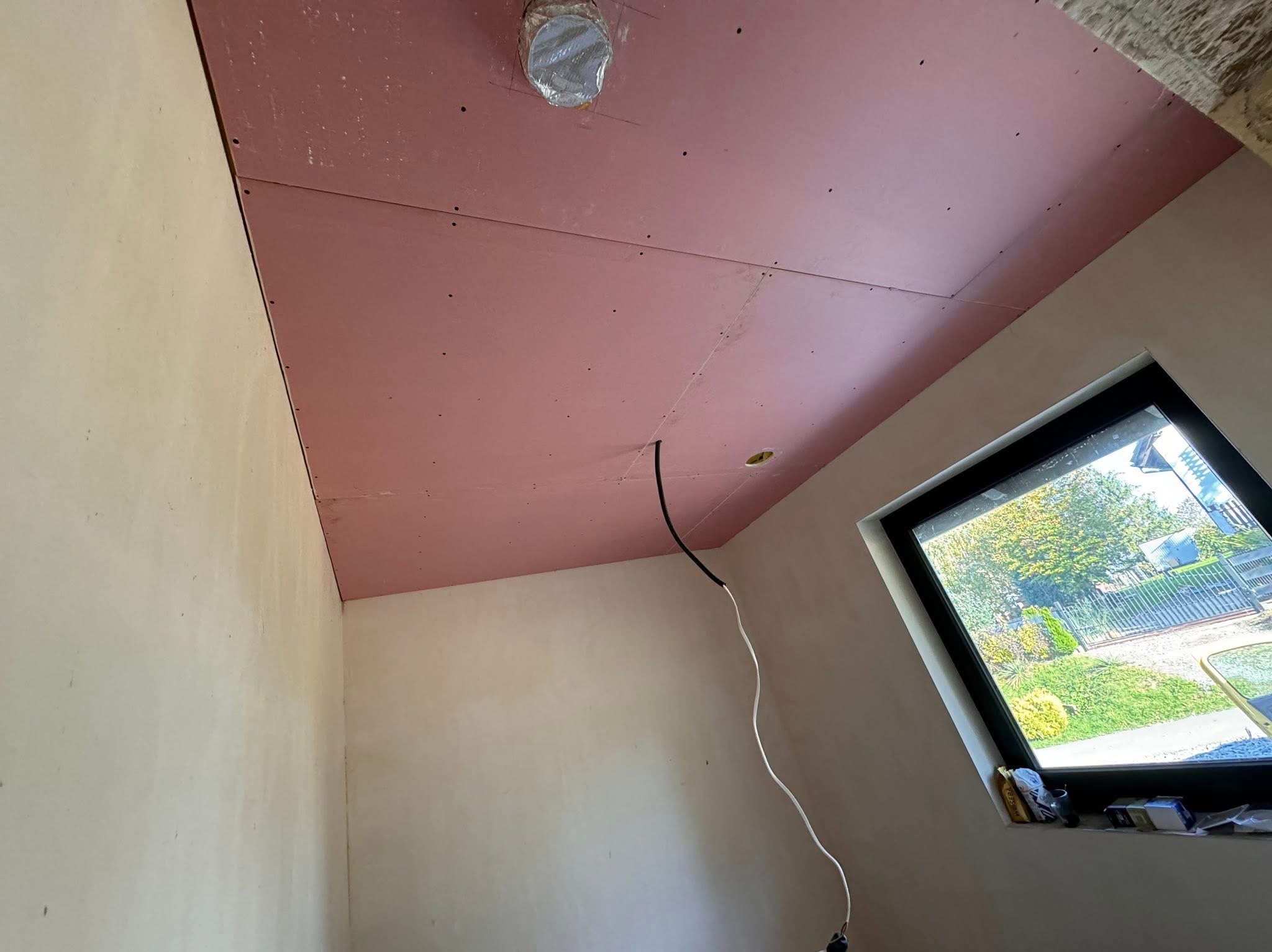 Różowa płyta karton-gips na suficie w trakcie montażu, widoczne kable i otwory na oświetlenie, surowe ściany i okno z widokiem na zewnątrz. Etap prac wykończeniowych.