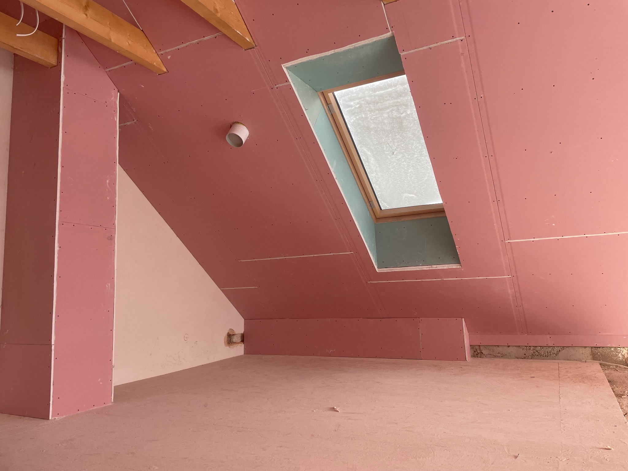 Poddasze z oknem dachowym, wykończone różową płytą kartonowo-gipsową. Widoczne drewniane elementy konstrukcyjne i fragment instalacji. Etap prac wykończeniowych.
