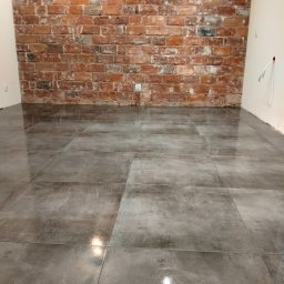 Mirosław Strzelec - Ułożone szare płytki podłogowe z połyskiem, na tle ściany z czerwonej cegły. Widoczny fragment białej ściany z gniazdkiem elektrycznym.