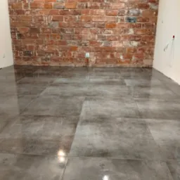 Ułożone szare płytki podłogowe z połyskiem, na tle ściany z czerwonej cegły. Widoczny fragment białej ściany z gniazdkiem elektrycznym.