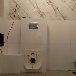 Mirosław Strzelec - Białe płytki imitujące marmur na ścianie łazienki, z widoczną instalacją podtynkową WC i częściowo ułożoną podłogą z płytek. Czerwony kabel zwisa z góry.