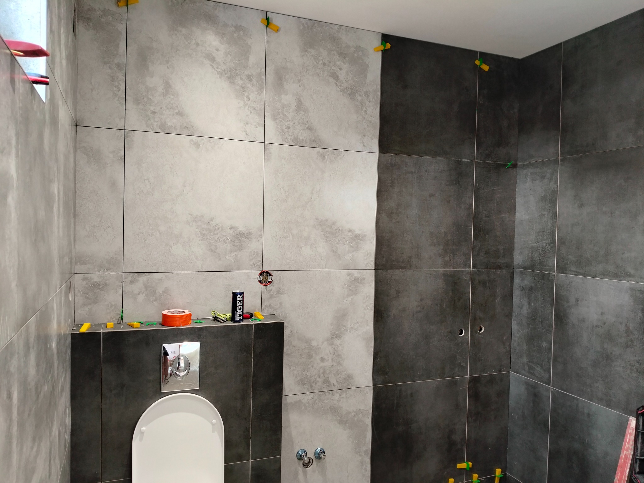 Ściana w łazience wyłożona szarymi płytkami z użyciem systemu poziomowania. Widoczne narzędzia, puszka i fragment toalety podtynkowej.