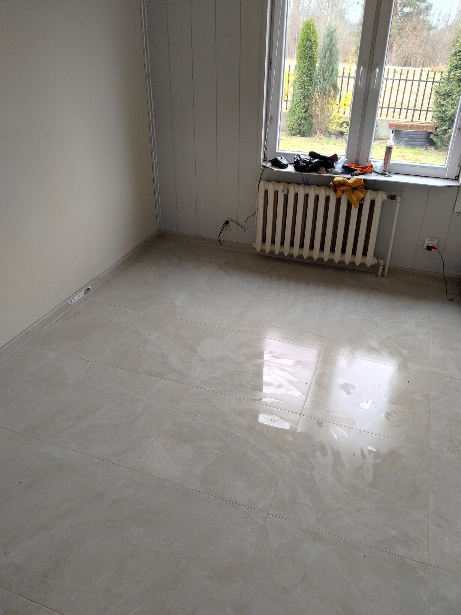 Jasne, błyszczące płytki podłogowe w pokoju z grzejnikiem pod oknem, ściany w panelach i gładzi. Widok na zewnątrz przez okno.