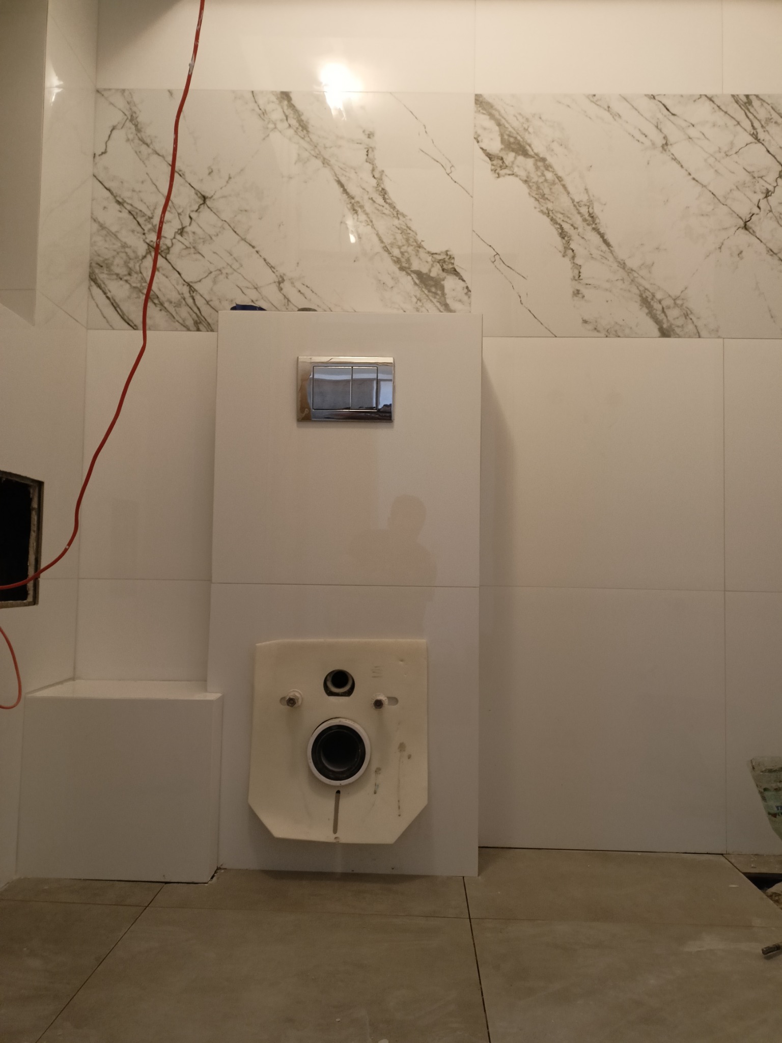 Białe płytki imitujące marmur na ścianie łazienki, z widoczną instalacją podtynkową WC i częściowo ułożoną podłogą z płytek. Czerwony kabel zwisa z góry.