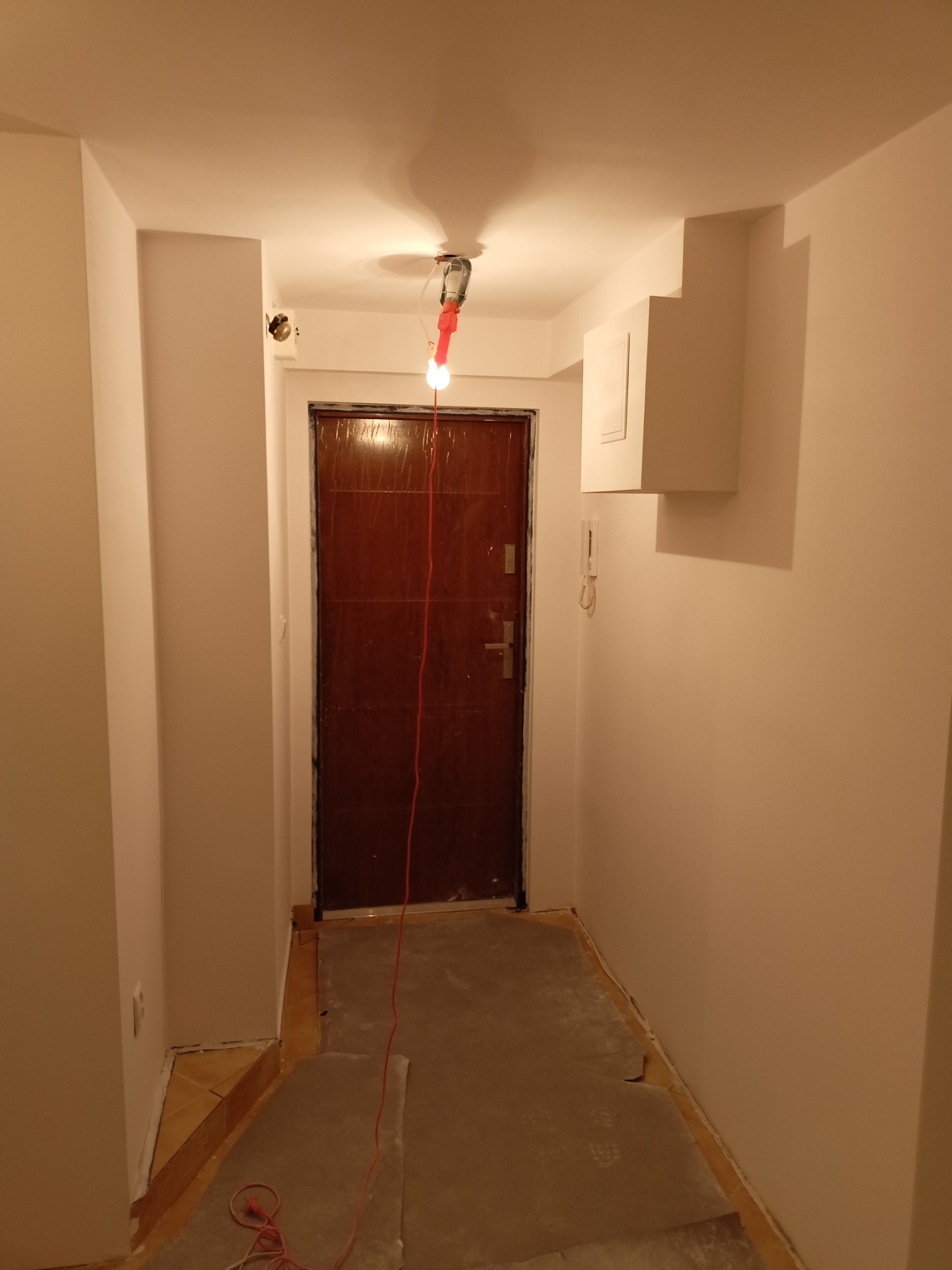 Korytarz po malowaniu na biało, widoczna brązowa stolarka drzwiowa i prowizoryczne oświetlenie. Podłoga zabezpieczona kartonem. Widoczny domofon.