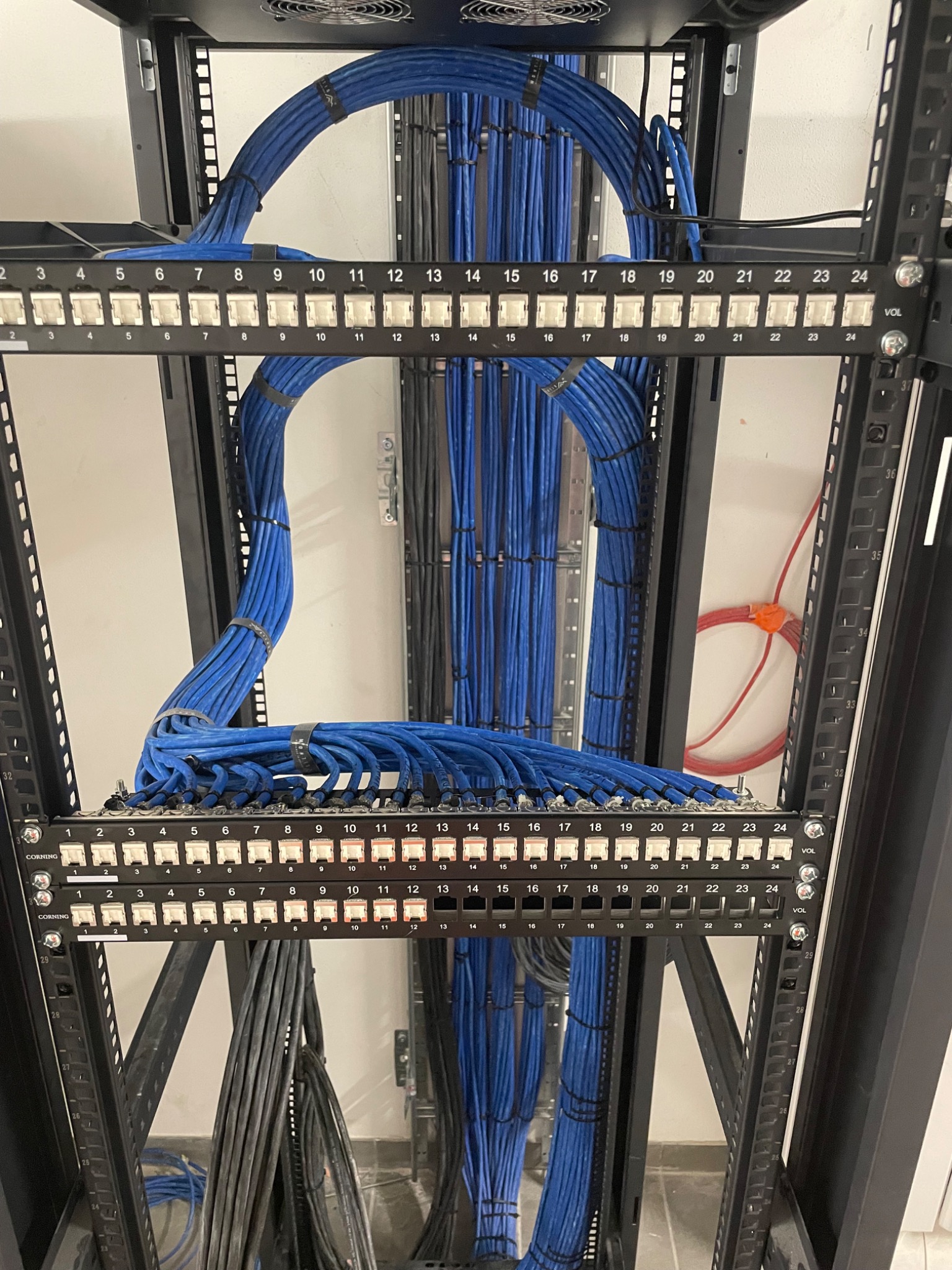 Panel krosowniczy z uporządkowanymi kablami sieciowymi w szafie rack, widok z bliska, etykiety portów, niebieskie kable spięte opaskami, wentylator w górnej części szafy.