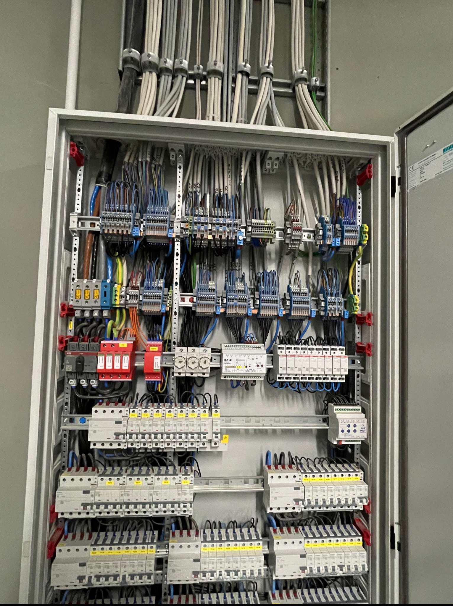 Otwarta szafa elektryczna z licznymi przewodami, bezpiecznikami i elementami sterującymi. Widoczne kable wchodzące do szafy od góry. Detaliczne ujęcie.