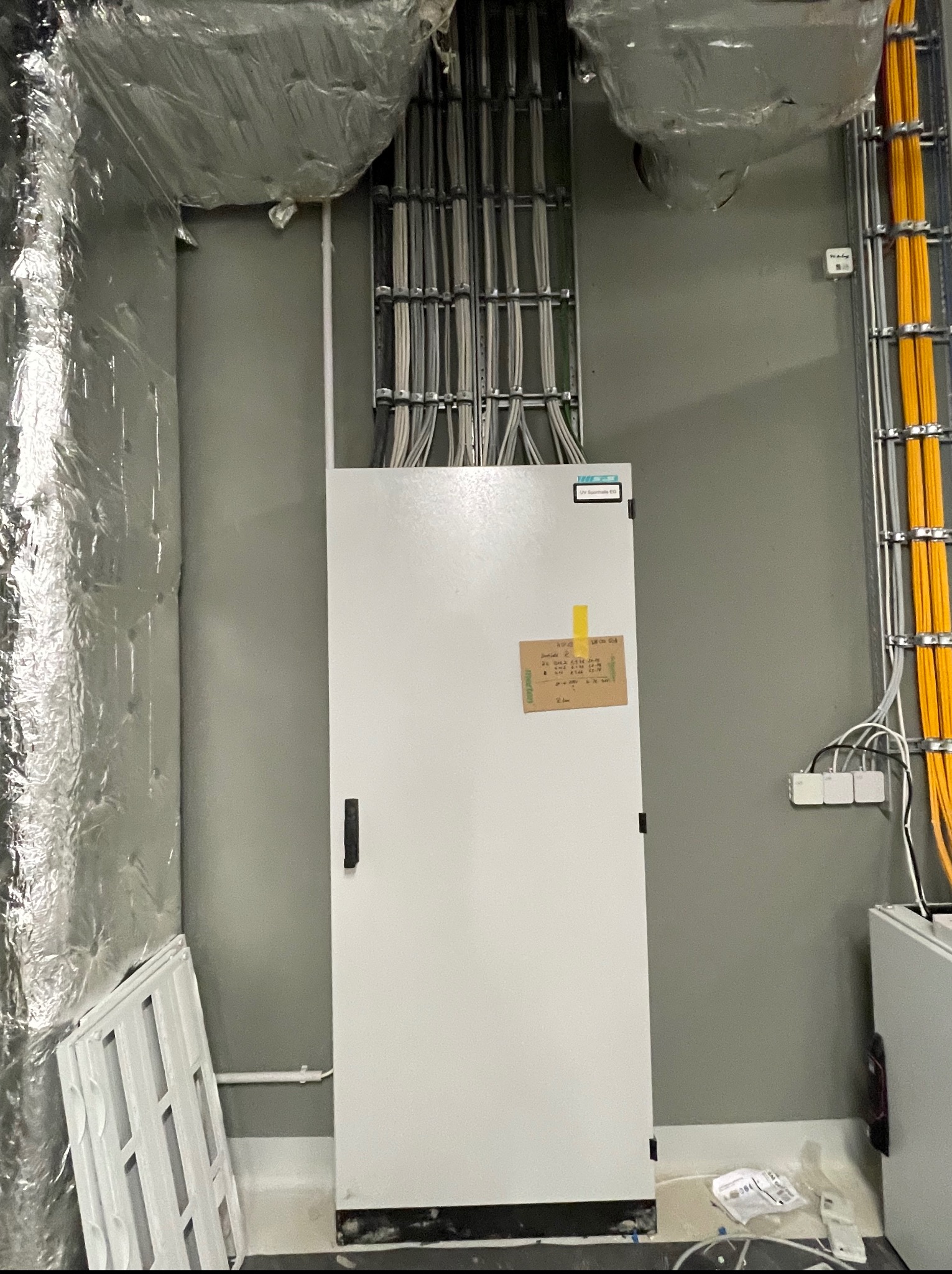 Biała szafa elektryczna z widocznymi kablami i przewodami, zamontowana w szarym pomieszczeniu. Na szafie naklejona kartka z informacjami. Widoczne elementy instalacji.