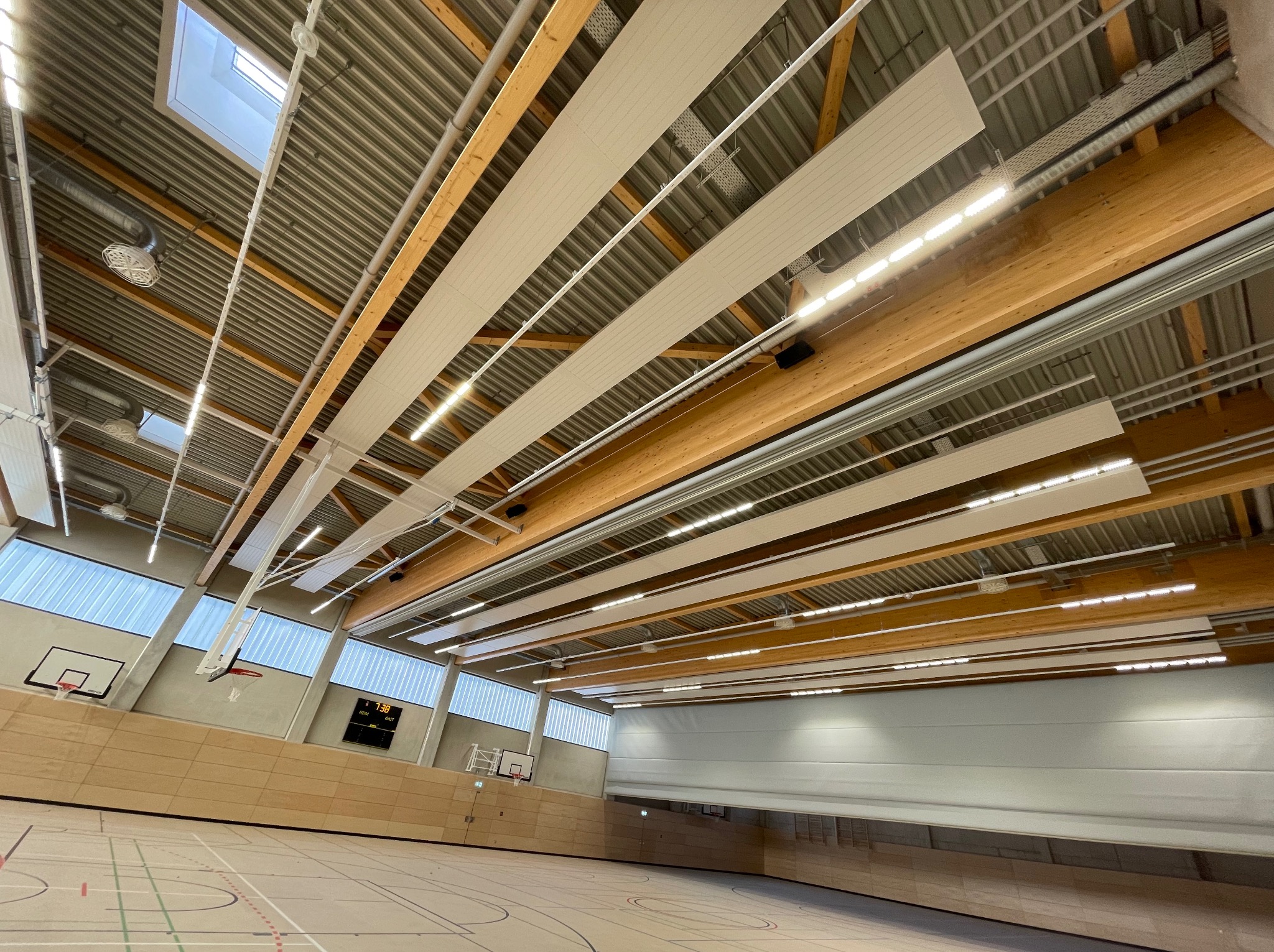 Oświetlenie LED w hali sportowej z drewnianą konstrukcją stropu i widocznymi elementami instalacji elektrycznej. Ujęcie z perspektywy parkietu.