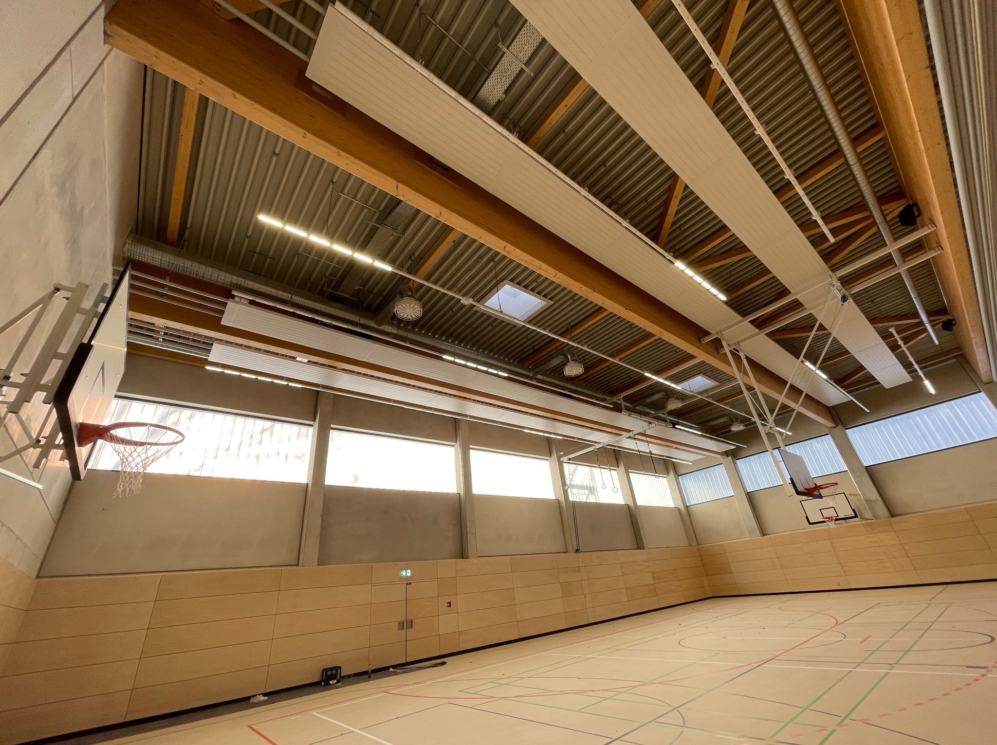 Wnętrze hali sportowej z drewnianą konstrukcją dachu, oświetleniem LED i parkietem z liniami boiska. Widoczne kosze do koszykówki i okna w górnej części ścian.