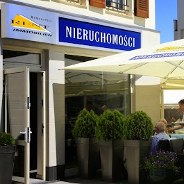 Fasada biura nieruchomości z szyldem 'KENT Immobilien NIERUCHOMOŚCI' i otwartymi drzwiami wejściowymi, widok z zewnątrz, z elementami kawiarni na pierwszym planie.