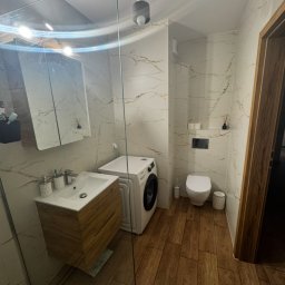 AF Investments Group - Nowoczesna łazienka z drewnianą szafką pod umywalką, białymi płytkami imitującymi marmur i pralką. Kabina prysznicowa ze szkła.