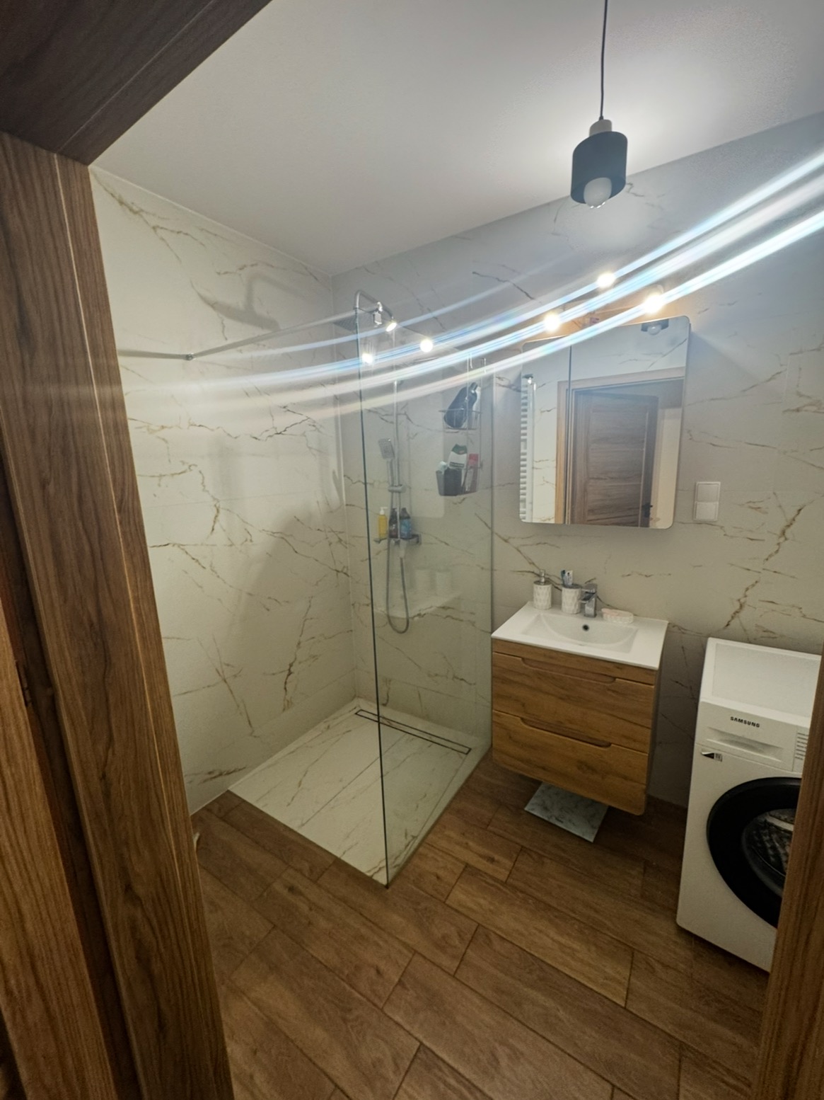 Nowoczesna łazienka po remoncie: kabina prysznicowa typu walk-in, drewniana szafka z umywalką, pralka Samsung, płytki imitujące marmur i drewno.