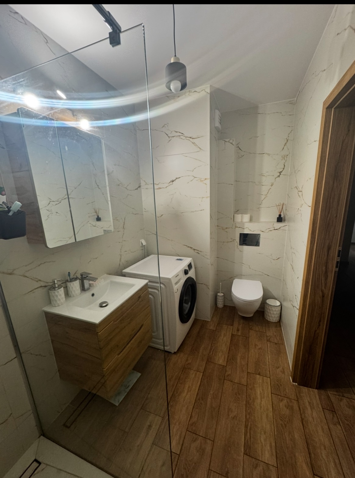 Nowoczesna łazienka z drewnianą szafką pod umywalką, białymi płytkami imitującymi marmur i pralką. Kabina prysznicowa ze szkła.