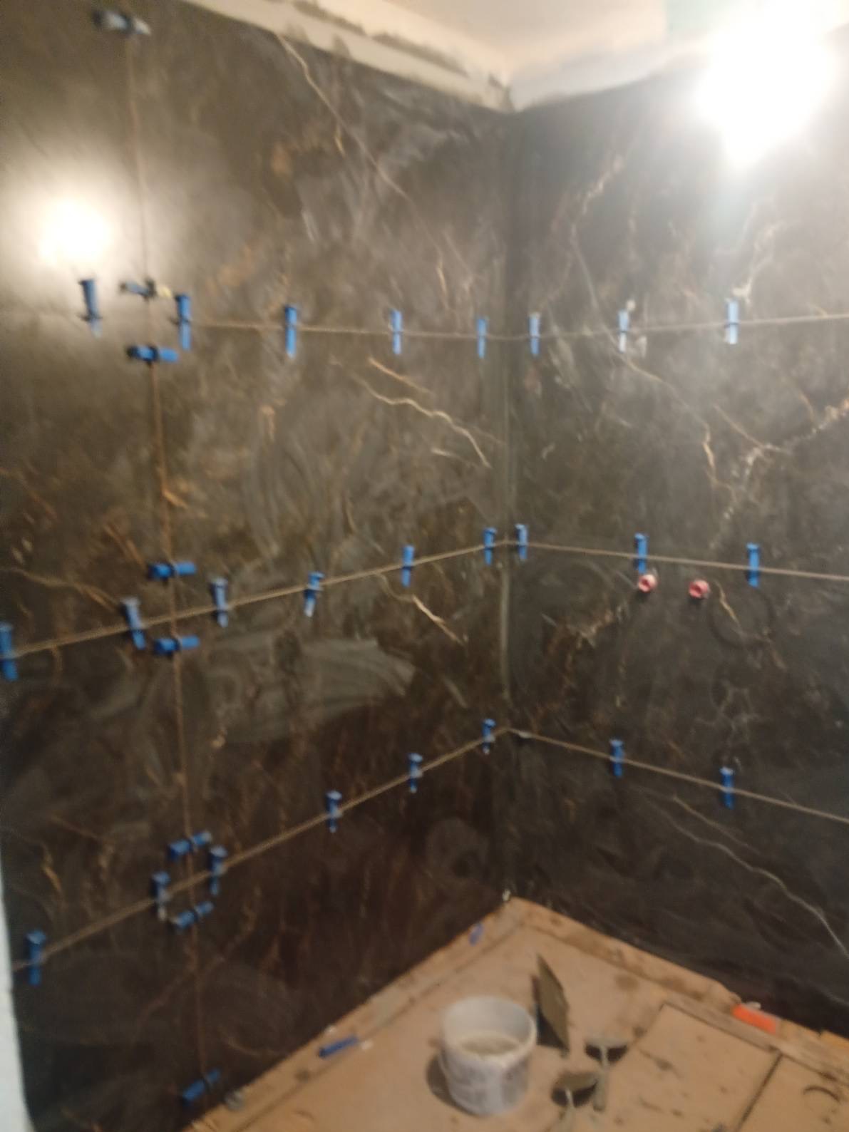 Ściana łazienki w trakcie układania płytek imitujących marmur, z użyciem poziomowania krzyżykami i sznurkiem. Widoczne narzędzia glazurnicze i wiadro z zaprawą.