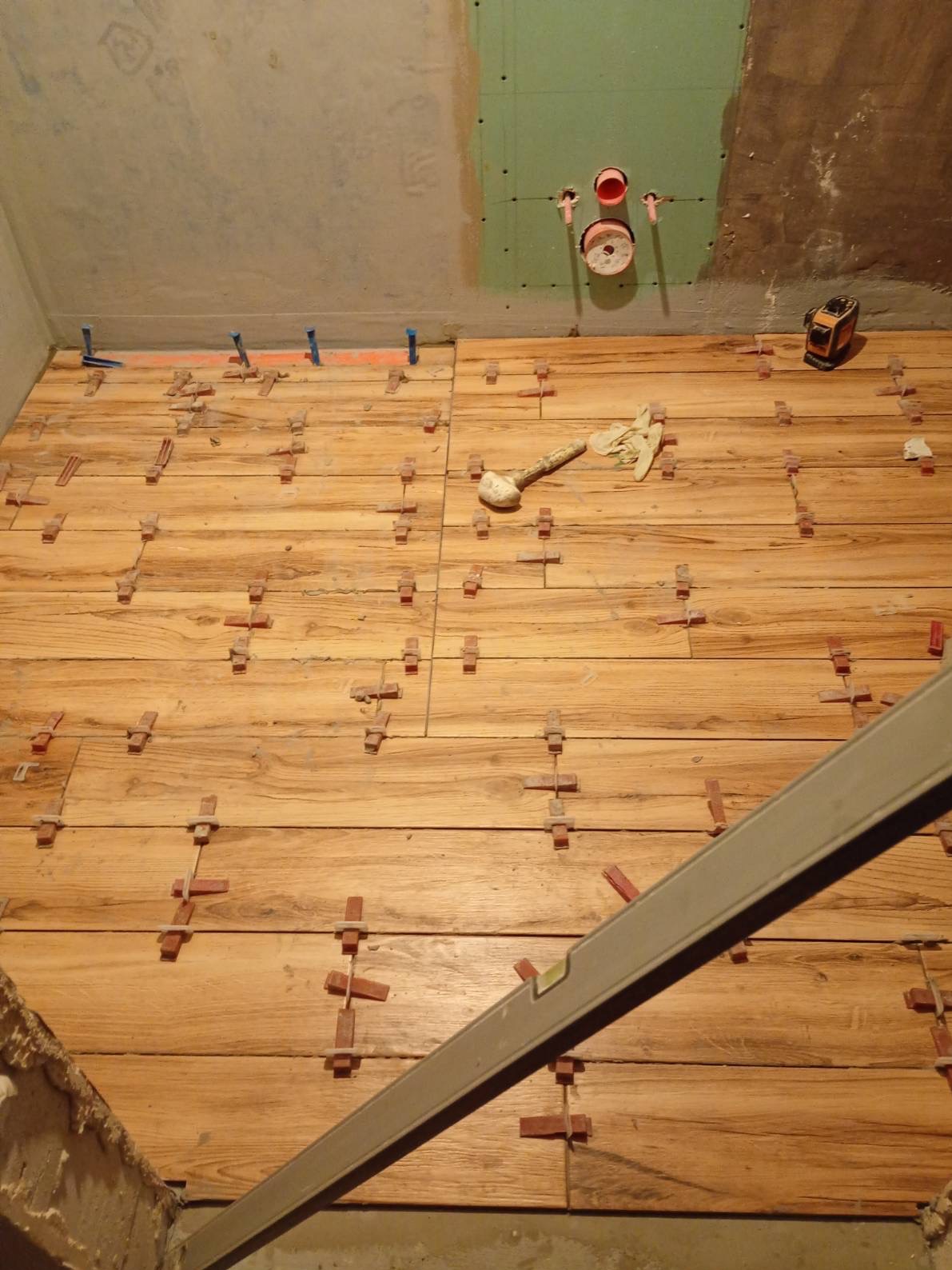 Układanie płytek drewnopodobnych na podłodze w łazience z użyciem systemu poziomowania, widoczny laser krzyżowy i poziomica.