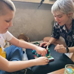 Katarzyna Marcińczak - Niania z krótkowłosą fryzurą i chłopiec układają klocki Lego na poduszce. Na stole stoi szklanka z napojem. Zabawa w domu.