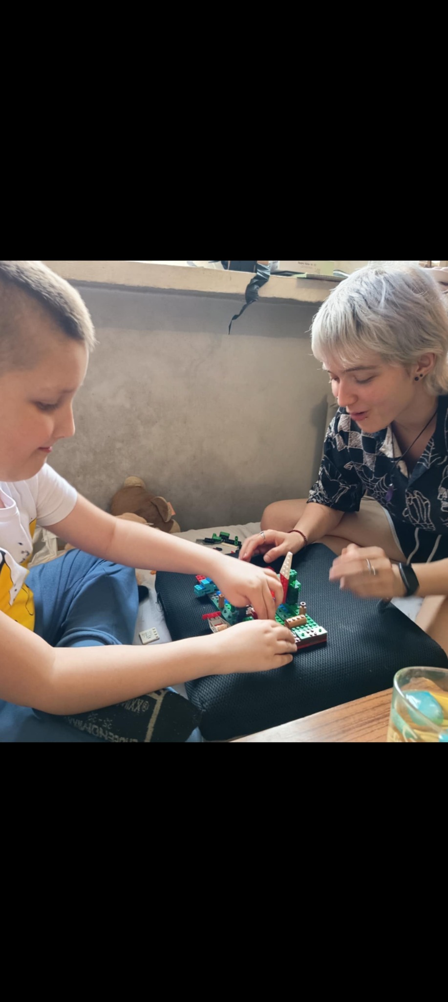 Niania z krótkowłosą fryzurą i chłopiec układają klocki Lego na poduszce. Na stole stoi szklanka z napojem. Zabawa w domu.