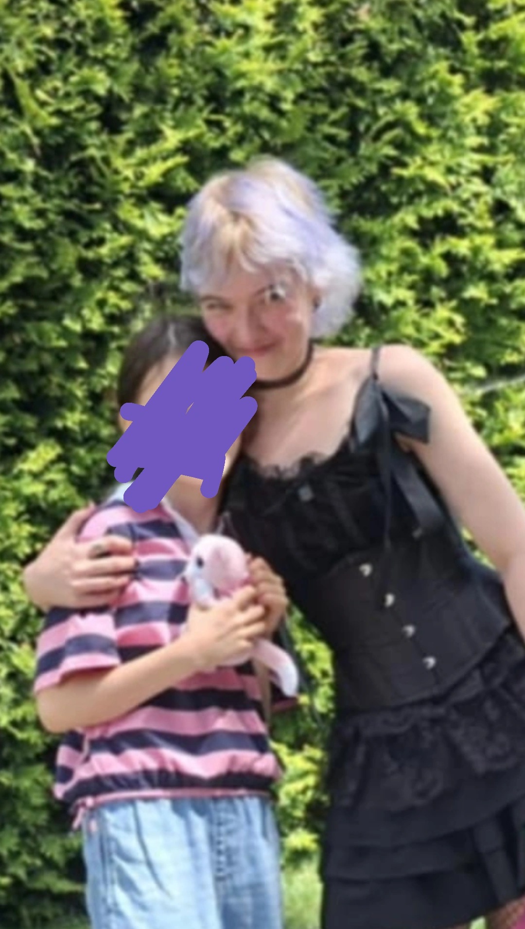 Opiekunka w czarnej sukience obejmuje dziewczynkę z pluszakiem na tle zielonego żywopłotu. Włosy opiekunki są krótkie i farbowane na blond z fioletowymi pasemkami.