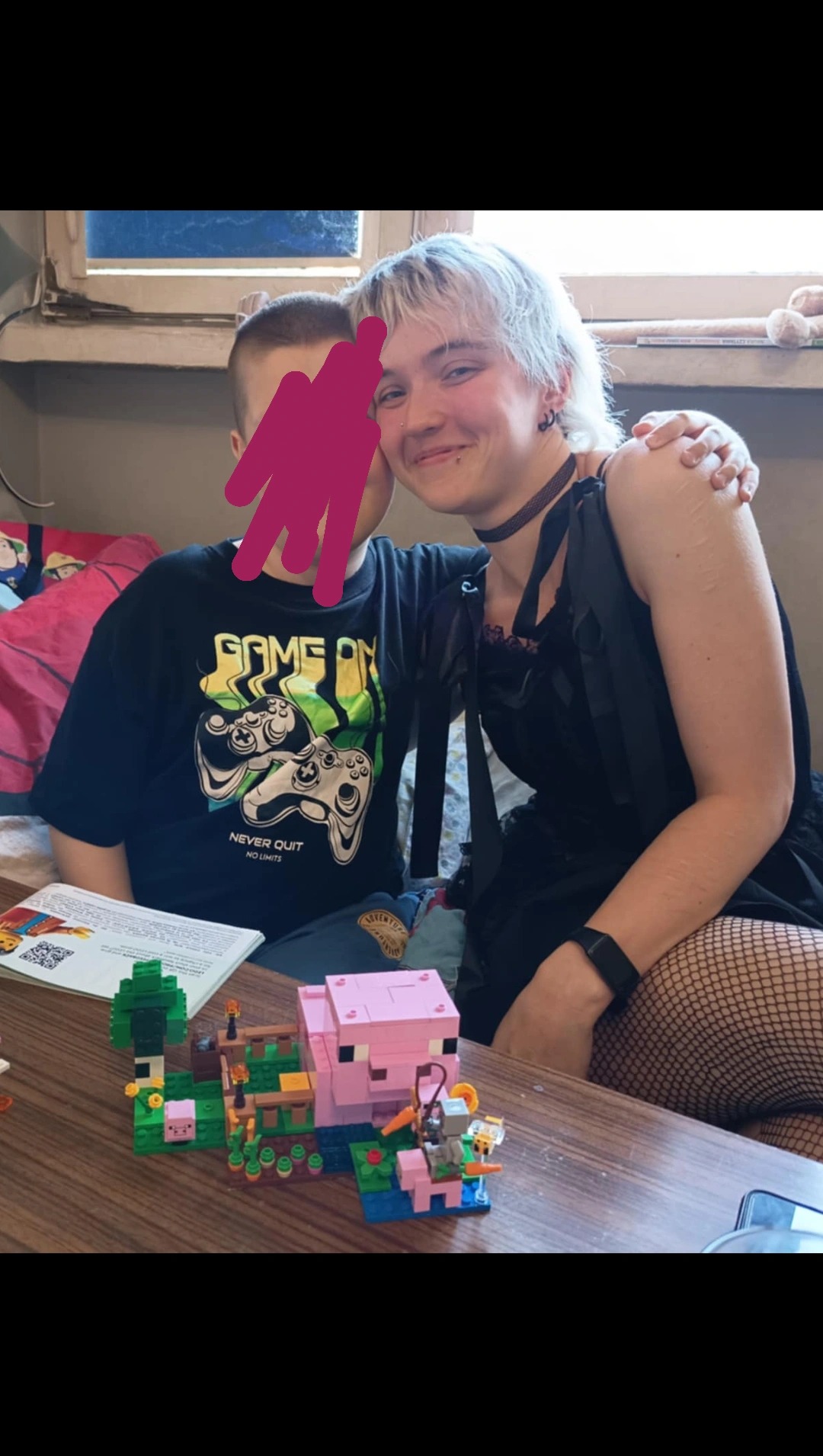 Uśmiechnięta kobieta z blond włosami obejmuje chłopca w koszulce z motywem gier. Na stole zestaw klocków Lego Minecraft. Domowe otoczenie, okno w tle.