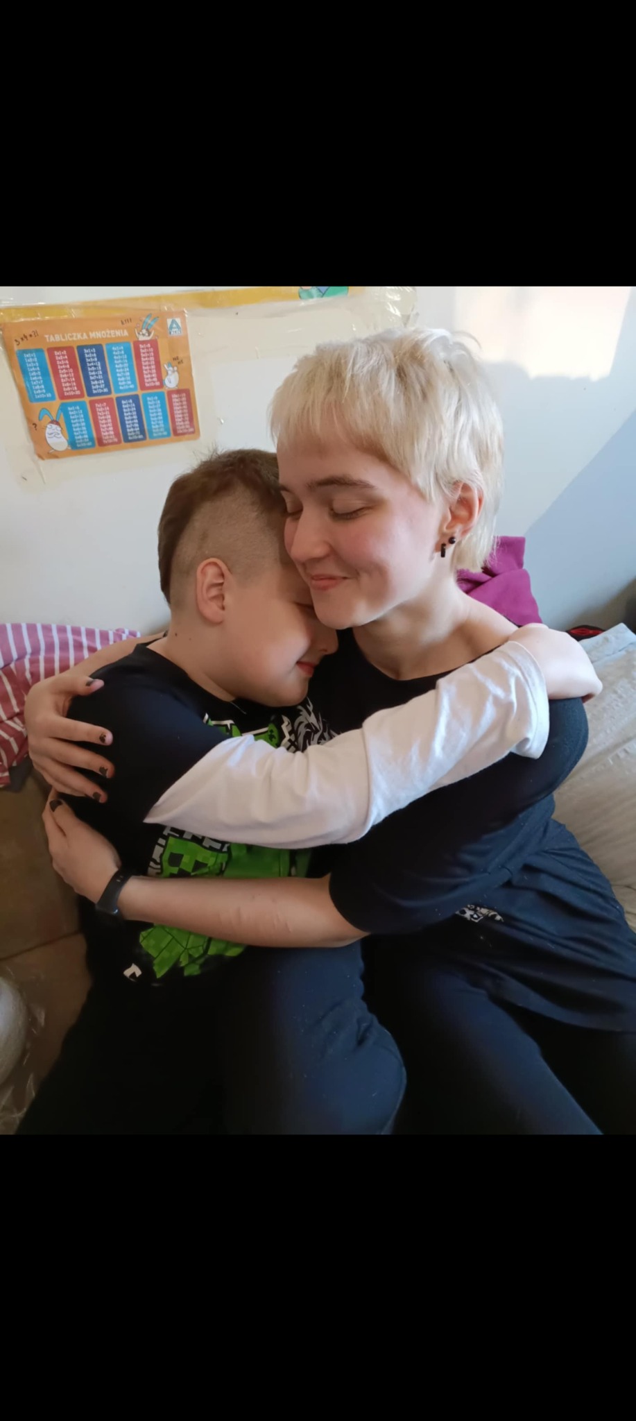 Młoda kobieta z krótkimi blond włosami czule obejmuje śpiącego chłopca w czarnej koszulce z Minecraftem. W tle widoczna tabliczka mnożenia na ścianie.