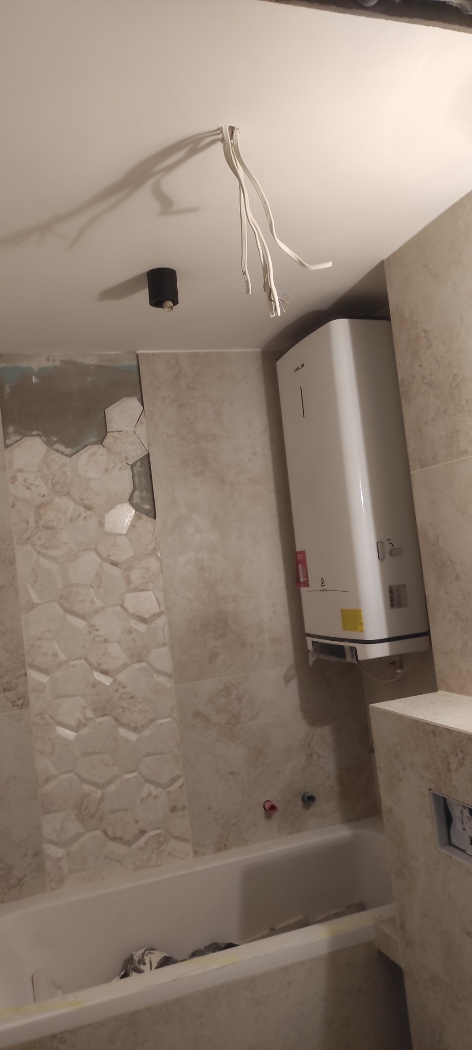 Remont łazienki w trakcie: położone płytki heksagonalne, biały bojler na ścianie, wystające kable. Widoczna wanna i niedokończone prace glazurnicze.