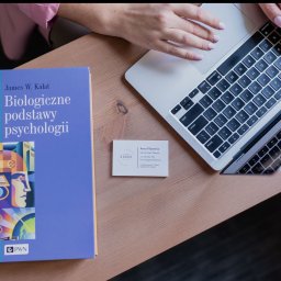 Gabinet Psychologiczny DOBRO Gliwice - Książka 'Biologiczne podstawy psychologii', wizytówka Anny Filipowicz i laptop na biurku. Kadr z góry, jasne oświetlenie, dłonie na klawiaturze.