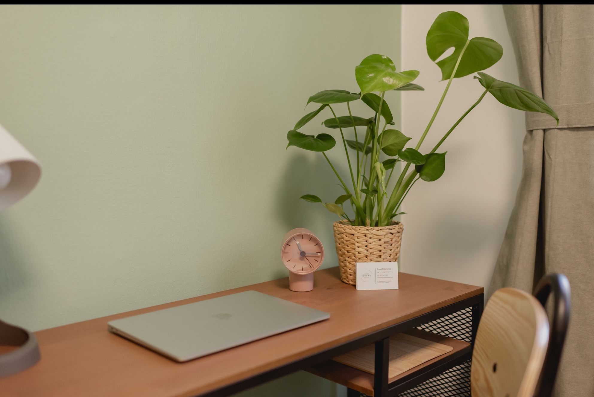 Biuro psychologa: minimalistyczne biurko z laptopem, zegarem, wizytówką i rośliną monstera na tle zielonej ściany i zasłony, tworzące spokojną atmosferę.