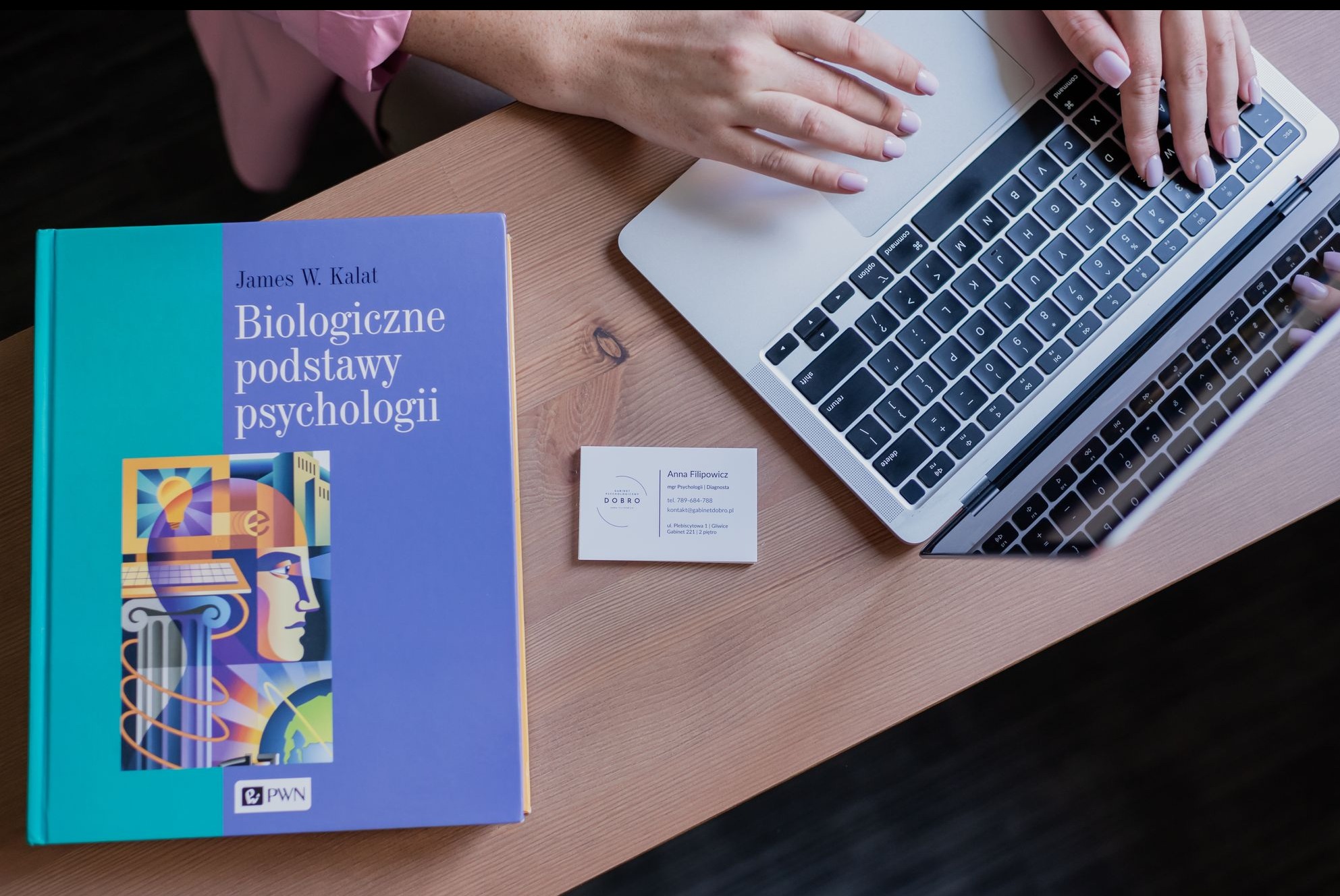 Książka 'Biologiczne podstawy psychologii', wizytówka Anny Filipowicz i laptop na biurku. Kadr z góry, jasne oświetlenie, dłonie na klawiaturze.