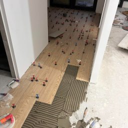 InnerLine Oleh Khomyn - Układanie drewnopodobnych płytek podłogowych z użyciem systemu poziomowania. Klej na podłodze, narzędzia glazurnicze. Widok z góry na fragment podłogi w trakcie prac.