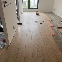 InnerLine Oleh Khomyn - Układanie podłogi z drewnopodobnych paneli w jasnym wnętrzu. Widoczne krzyżyki dystansowe, wiadro z klejem i fragment surowej podłogi. Częściowo położona podłoga.