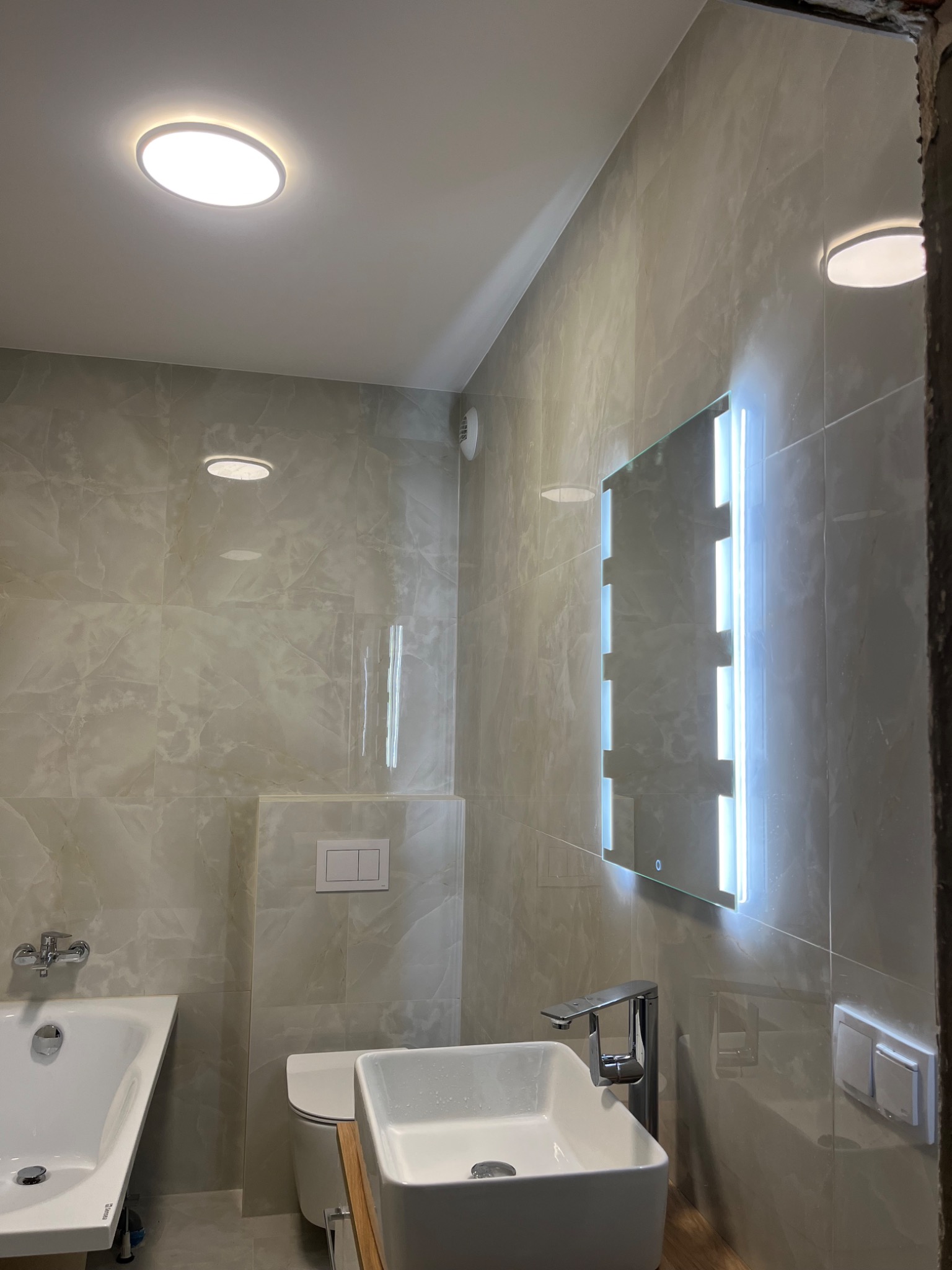 Jasna łazienka z beżowymi płytkami imitującymi marmur, podświetlane lustro, nowoczesna umywalka i bateria. Widok na wannę i toaletę. Oświetlenie sufitowe.