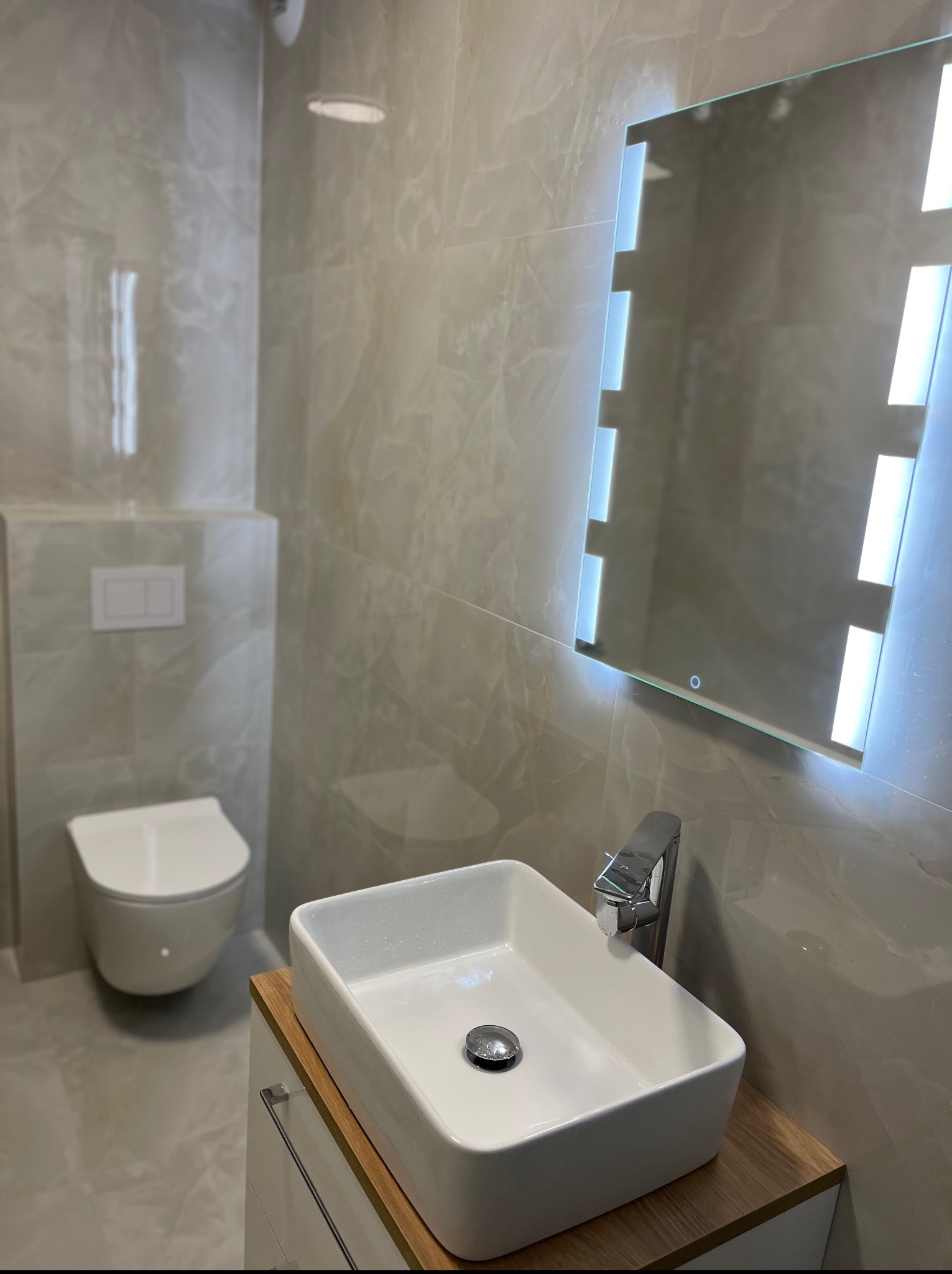 Nowoczesna łazienka z podwieszaną toaletą, umywalką nablatową na drewnianym blacie i lustrem z podświetleniem LED, wykończona płytkami imitującymi marmur.