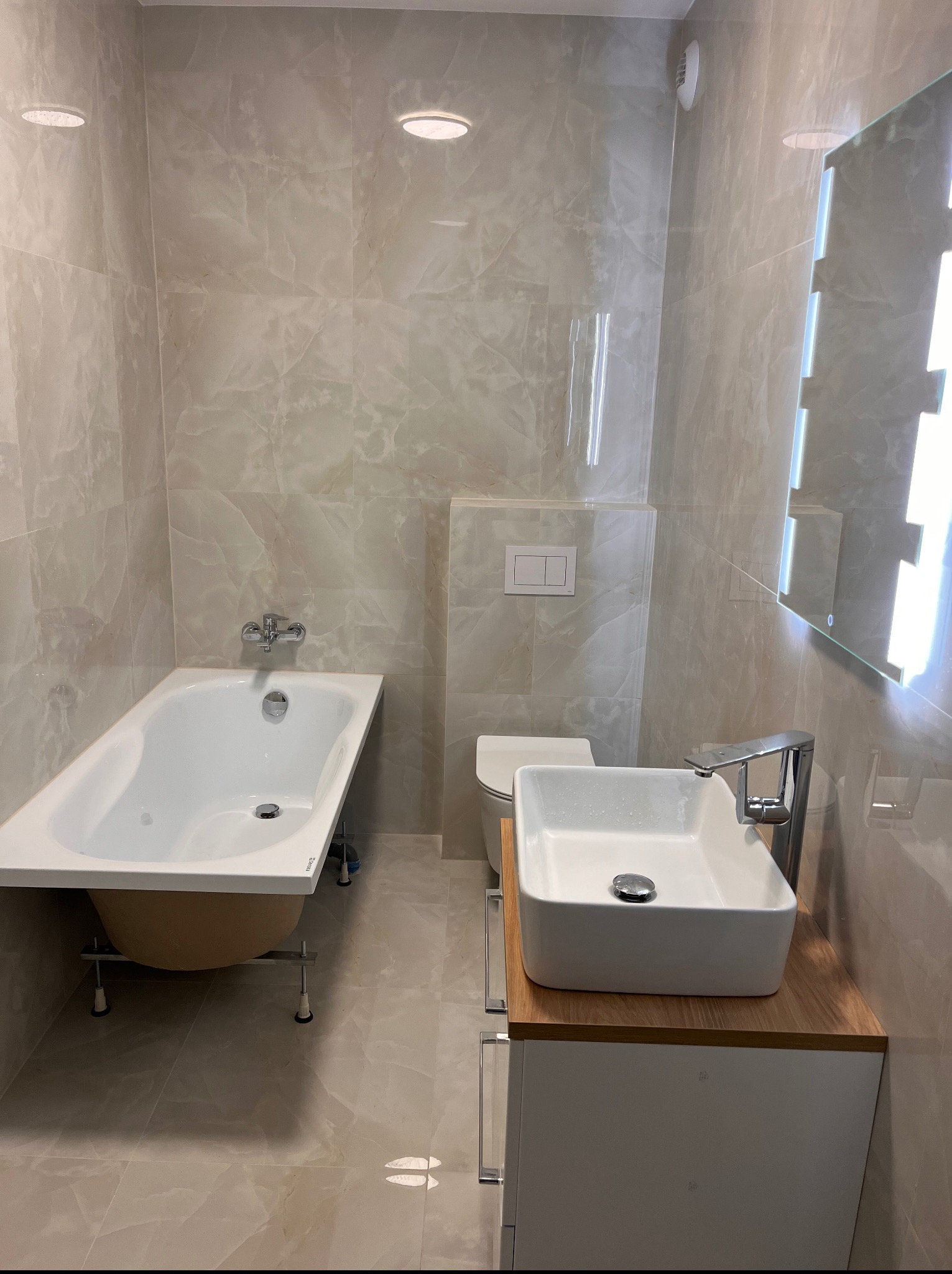 Jasna łazienka po remoncie: wanna na nóżkach, umywalka nablatowa z szafką, toaleta w zabudowie, płytki imitujące marmur, lustro z podświetleniem LED.