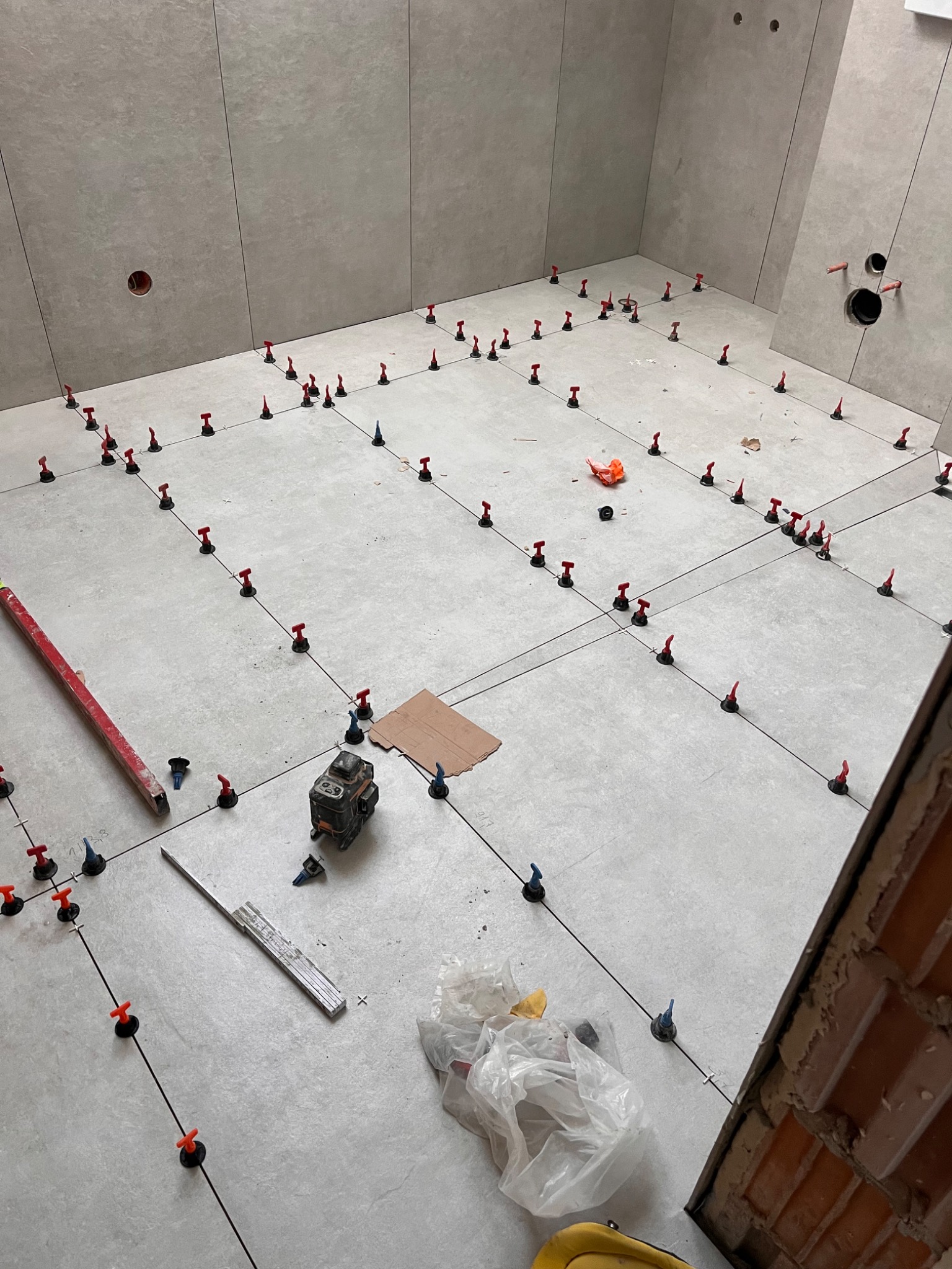 Jasnoszare płytki podłogowe z systemem poziomowania w trakcie układania. Widoczny laser krzyżowy, poziomica i fragment ściany z cegły.