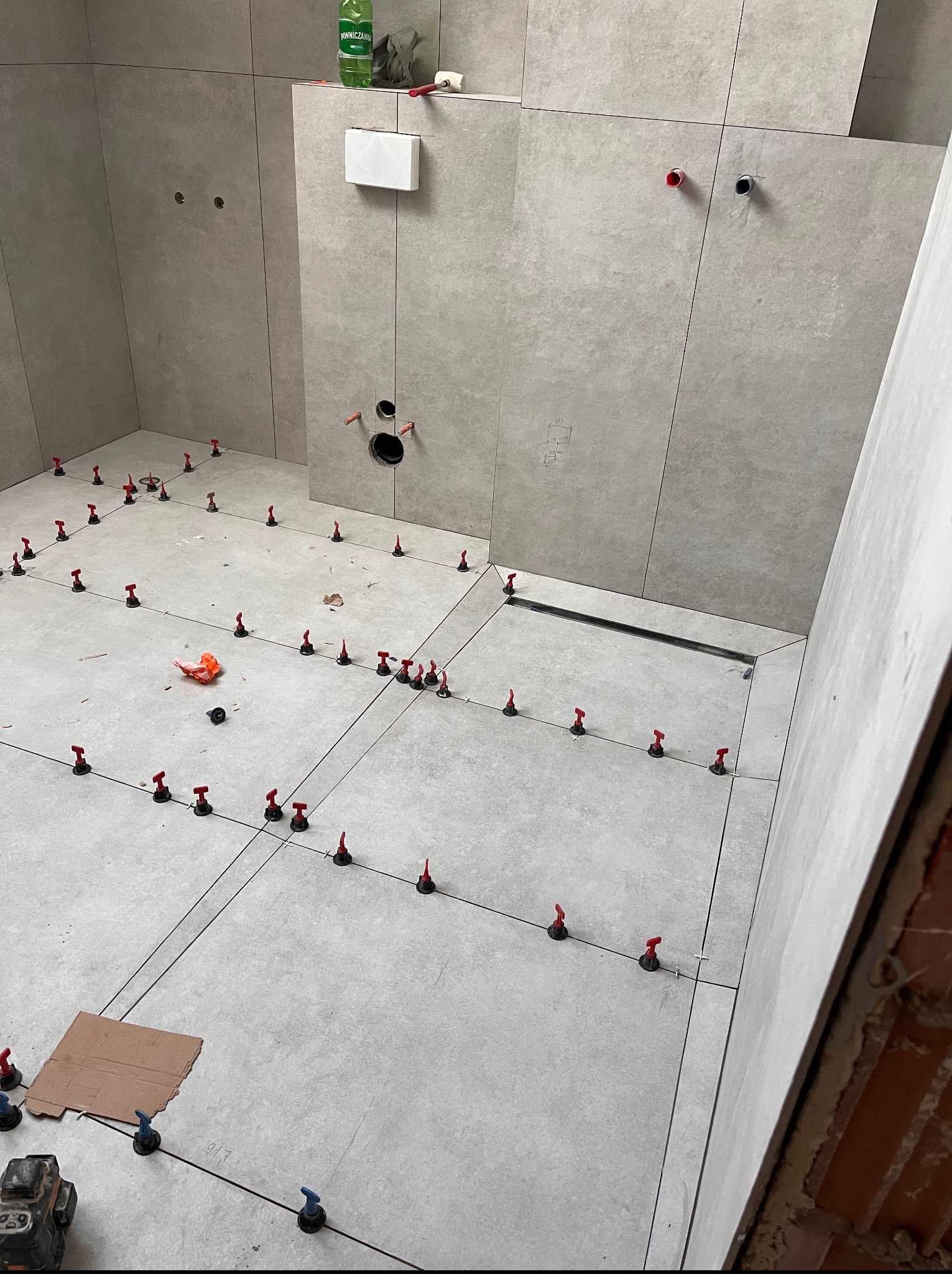 Położona, jasnoszara glazura w łazience, z użyciem systemu poziomowania płytek. Widoczne narzędzia, odpływ liniowy i przygotowane otwory na armaturę.