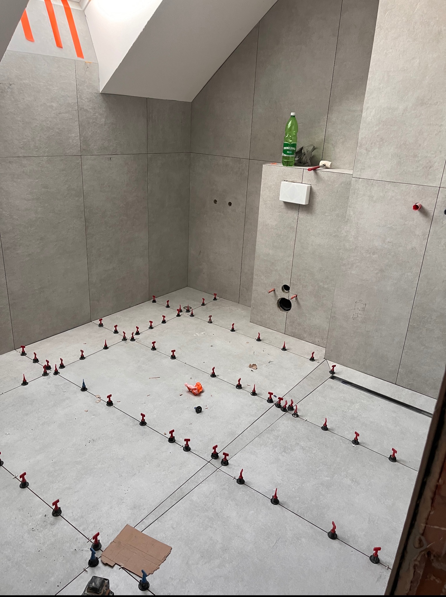 Szare płytki podłogowe i ścienne w trakcie układania w łazience na poddaszu. System poziomowania płytek z czerwonymi klipsami i czarnymi krzyżykami.