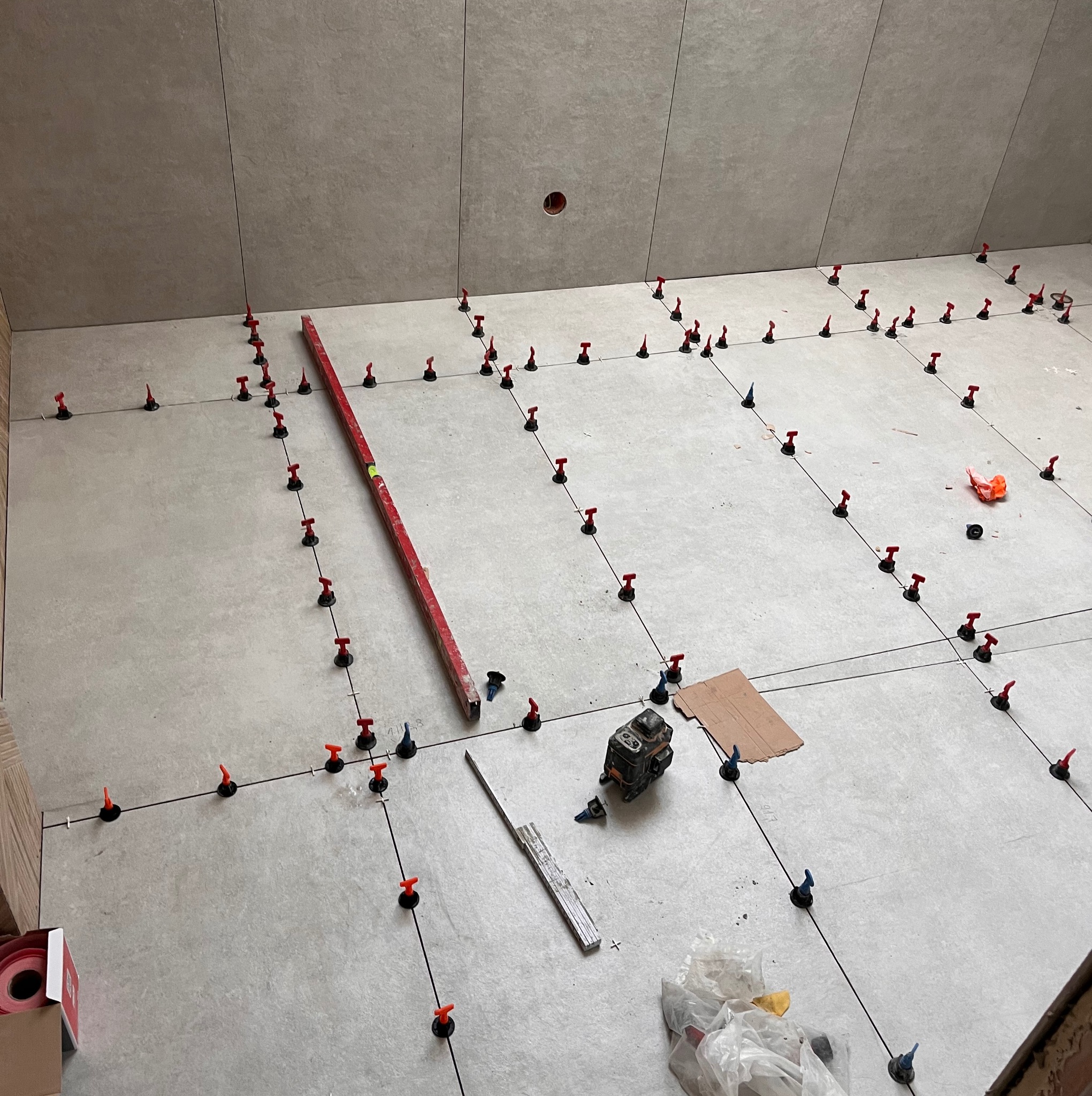 Szare płytki podłogowe w trakcie układania, z użyciem systemu poziomowania. Widoczny laser krzyżowy, poziomica i narzędzia glazurnicze.