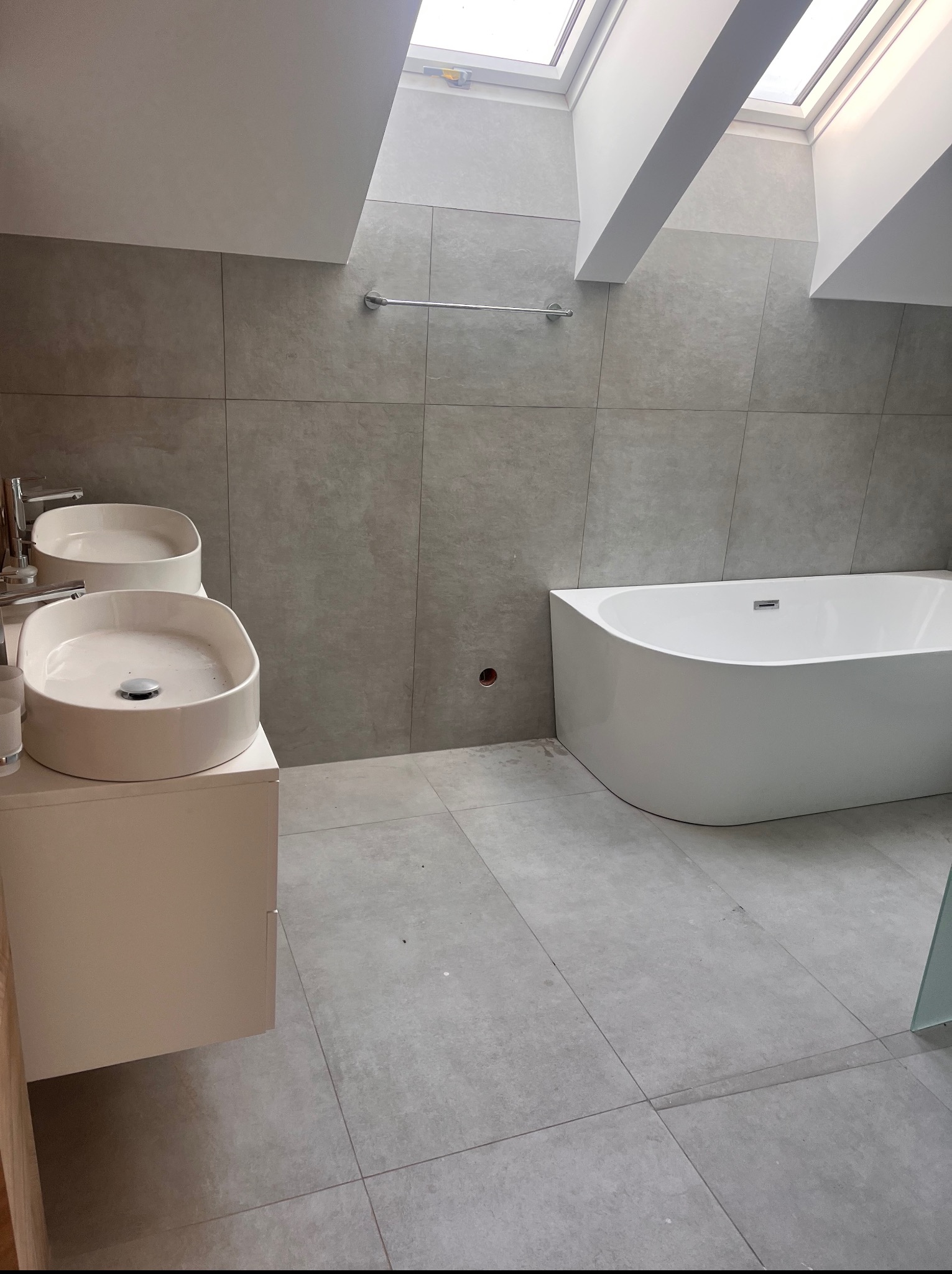 Jasna łazienka na poddaszu z dwoma umywalkami na szafce i białą wanną. Szare płytki na ścianach i podłodze, okno dachowe.