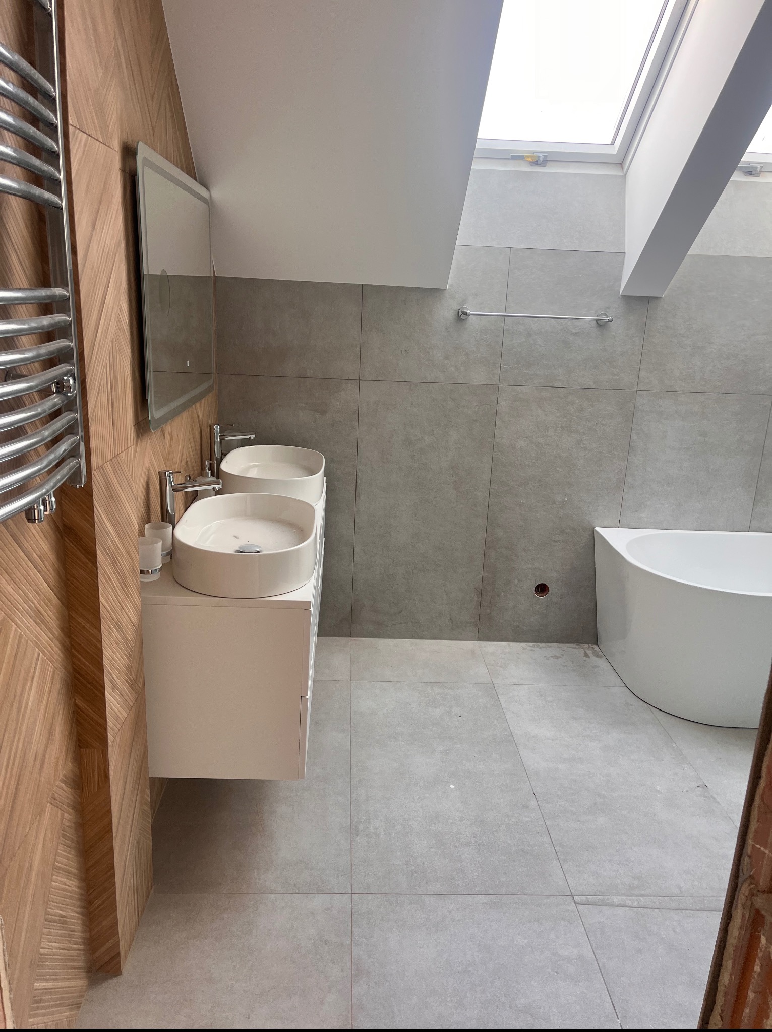 Nowoczesna łazienka z podwójną umywalką nablatową, szarą glazurą i drewnianą ścianą, oświetlona przez okno dachowe.