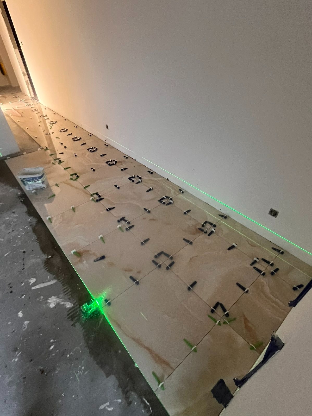 Układanie beżowych płytek z użyciem lasera i krzyżyków dystansowych. Widoczny klej i narzędzia. Prace glazurnicze w toku, precyzyjne poziomowanie.